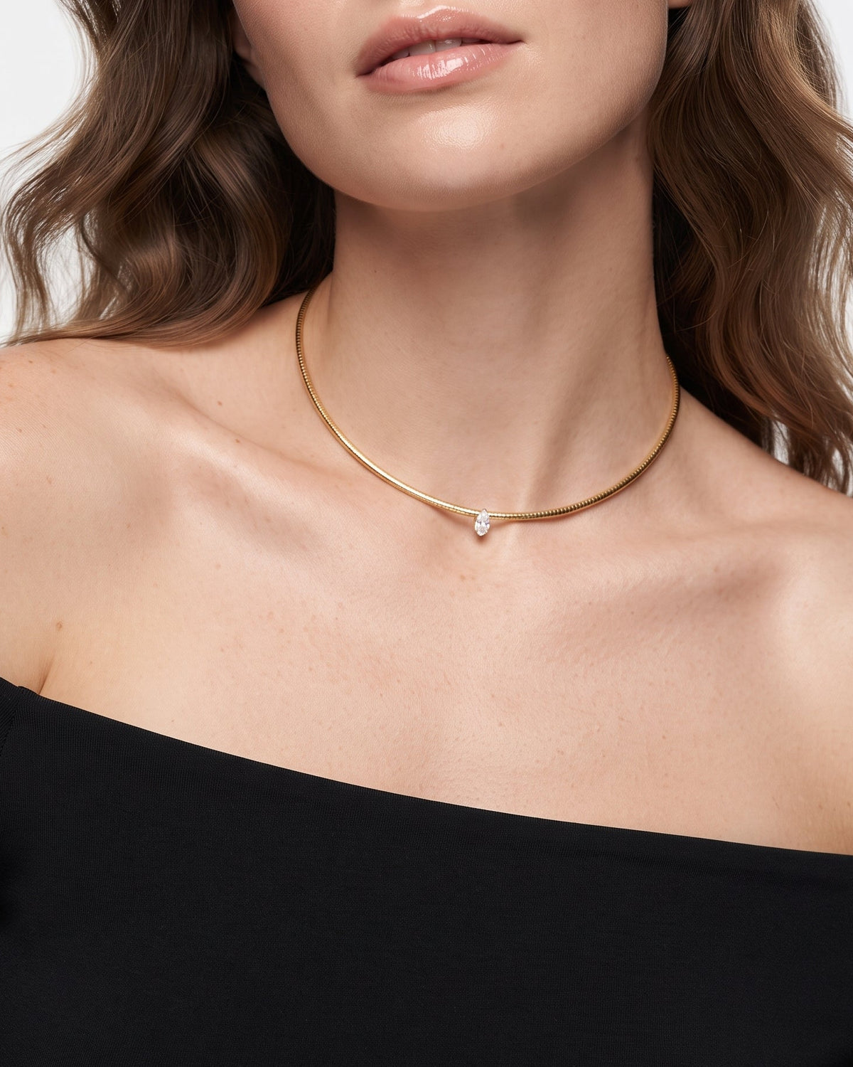 A close-up of a woman wearing a delicate gold chain necklace with a small white pearl pendant, paired with a black off-the-shoulder top against a light gray background.
