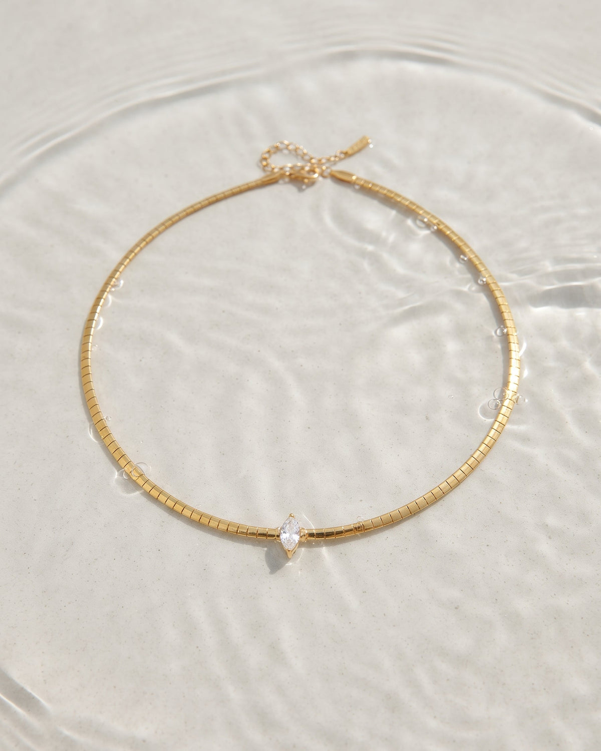 A delicate gold chain necklace with a small clear teardrop gemstone pendant, resting on a textured white surface with soft water ripples.