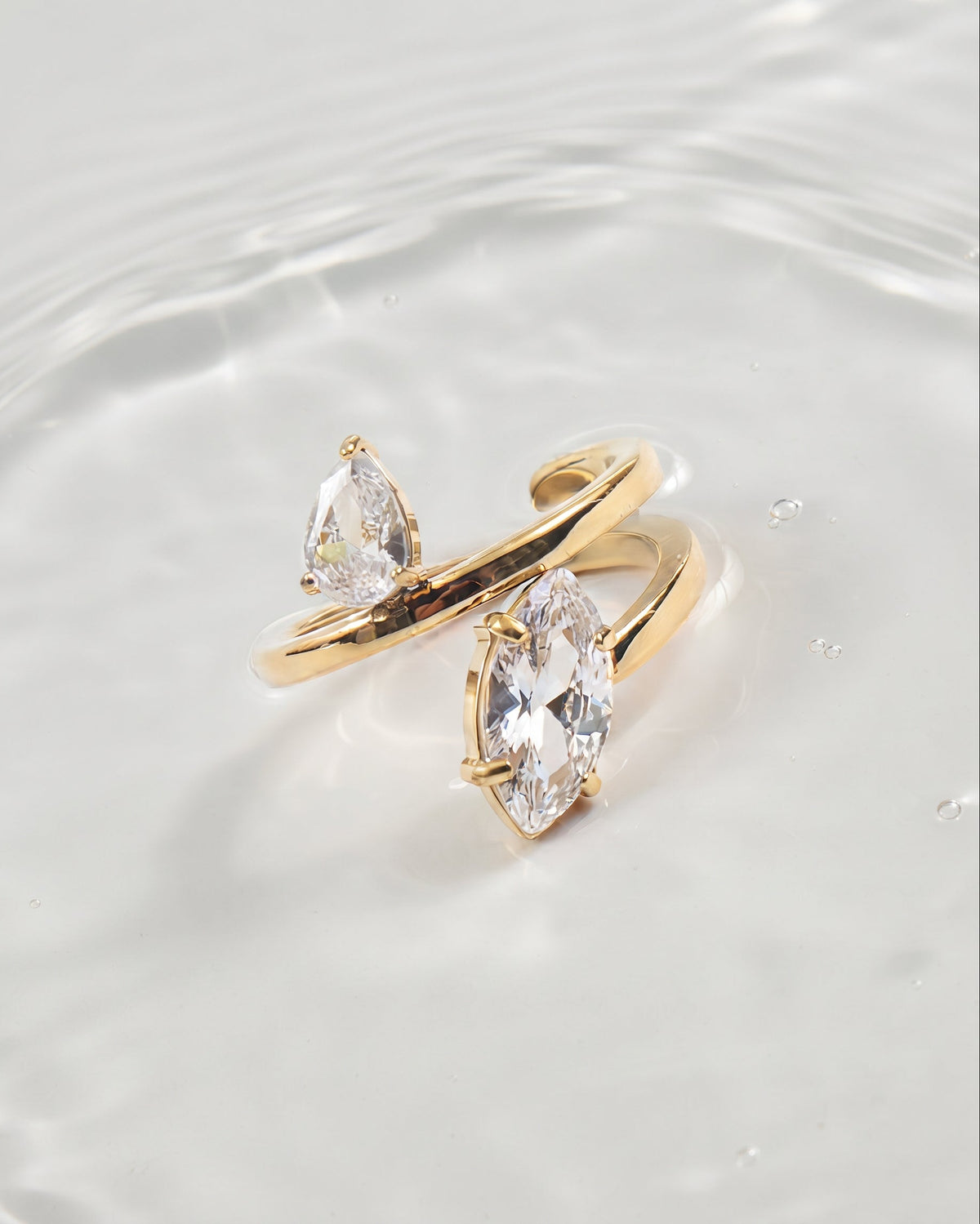 Two gold rings with marquise-cut clear gemstones, displayed on a glossy white surface with soft water ripples.