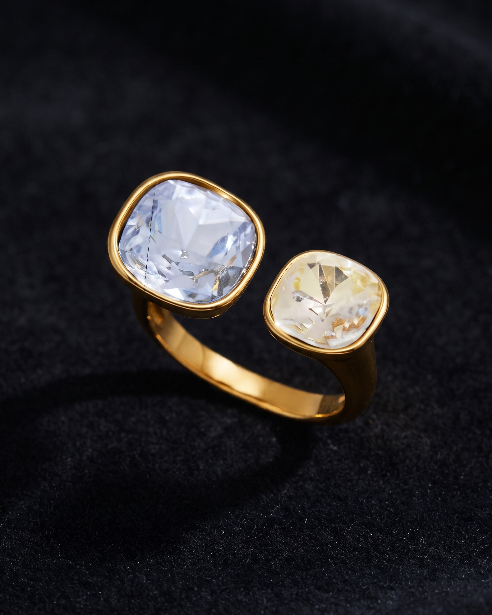 A gold ring with two cushion-cut gemstones, one large clear stone and one smaller yellow stone, set against a dark textured fabric background.
