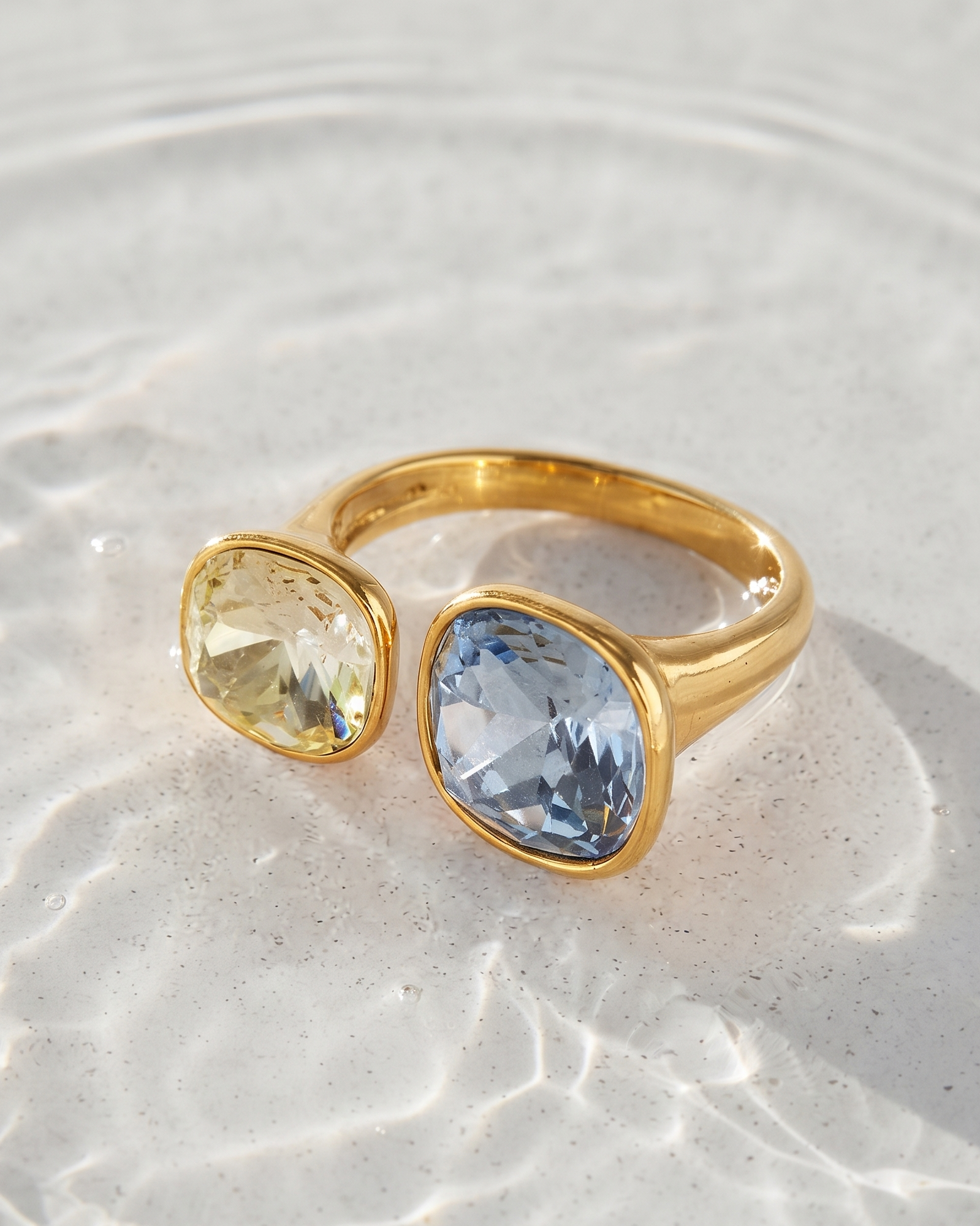 A gold ring with two cushion-cut gemstones, one pale yellow and one light blue, resting on a textured light gray surface with soft water ripples.