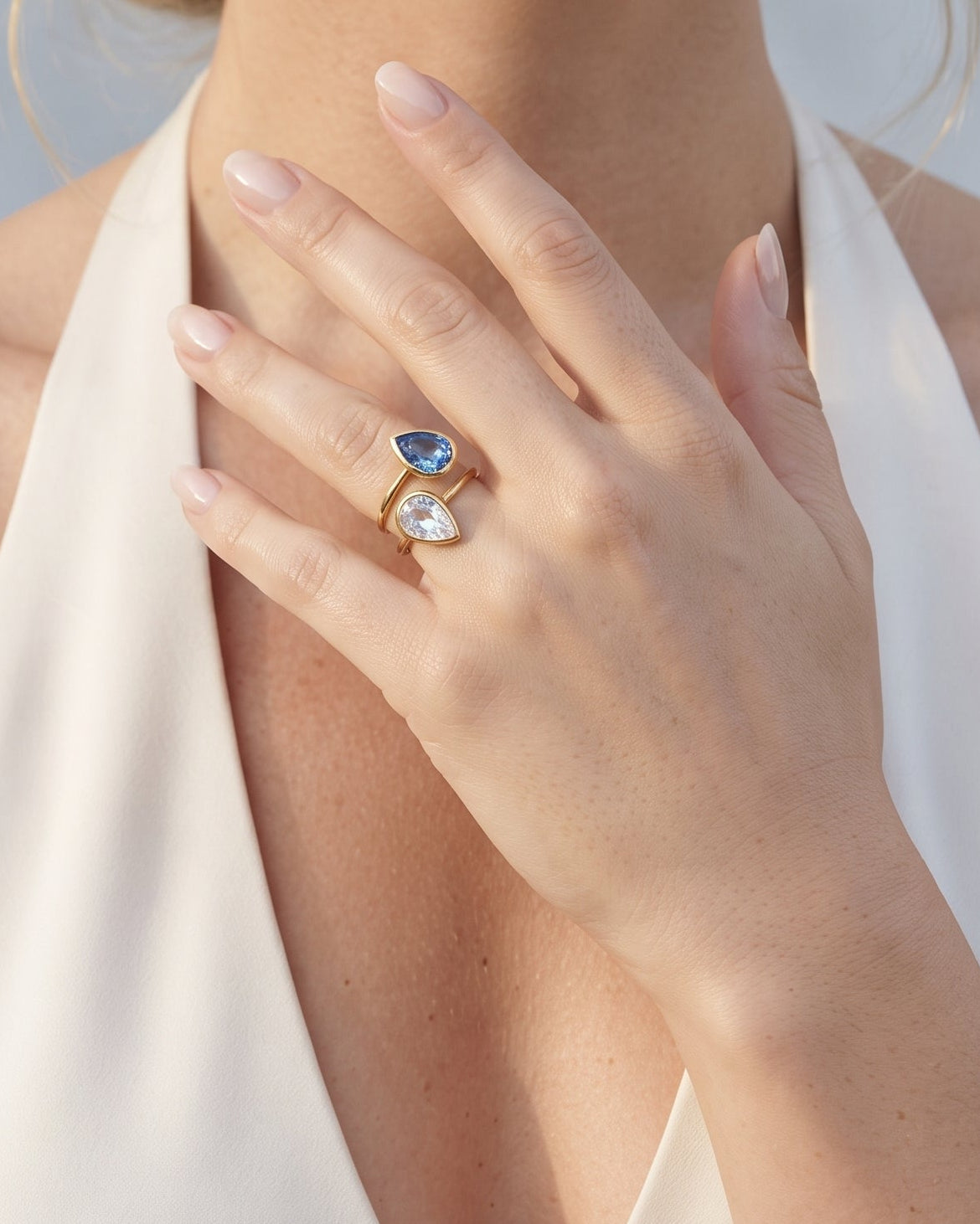 A close-up lifestyle shot of a woman wearing a gold ring with a marquise-cut blue gemstone and a clear heart-shaped gemstone, set against a soft gray background.