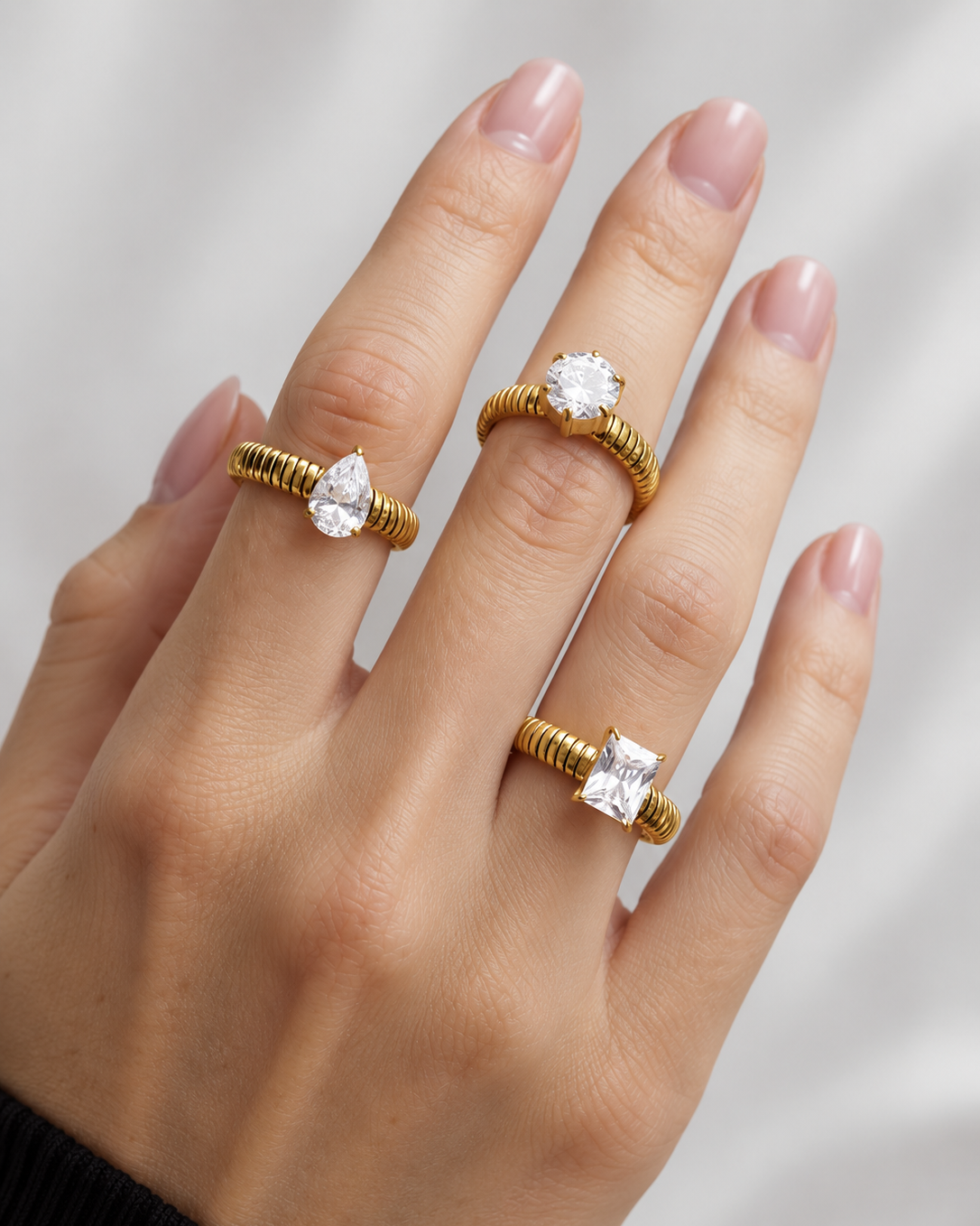 A close-up of a hand wearing three gold rings with clear gemstones in different shapes, set against a soft light gray background.