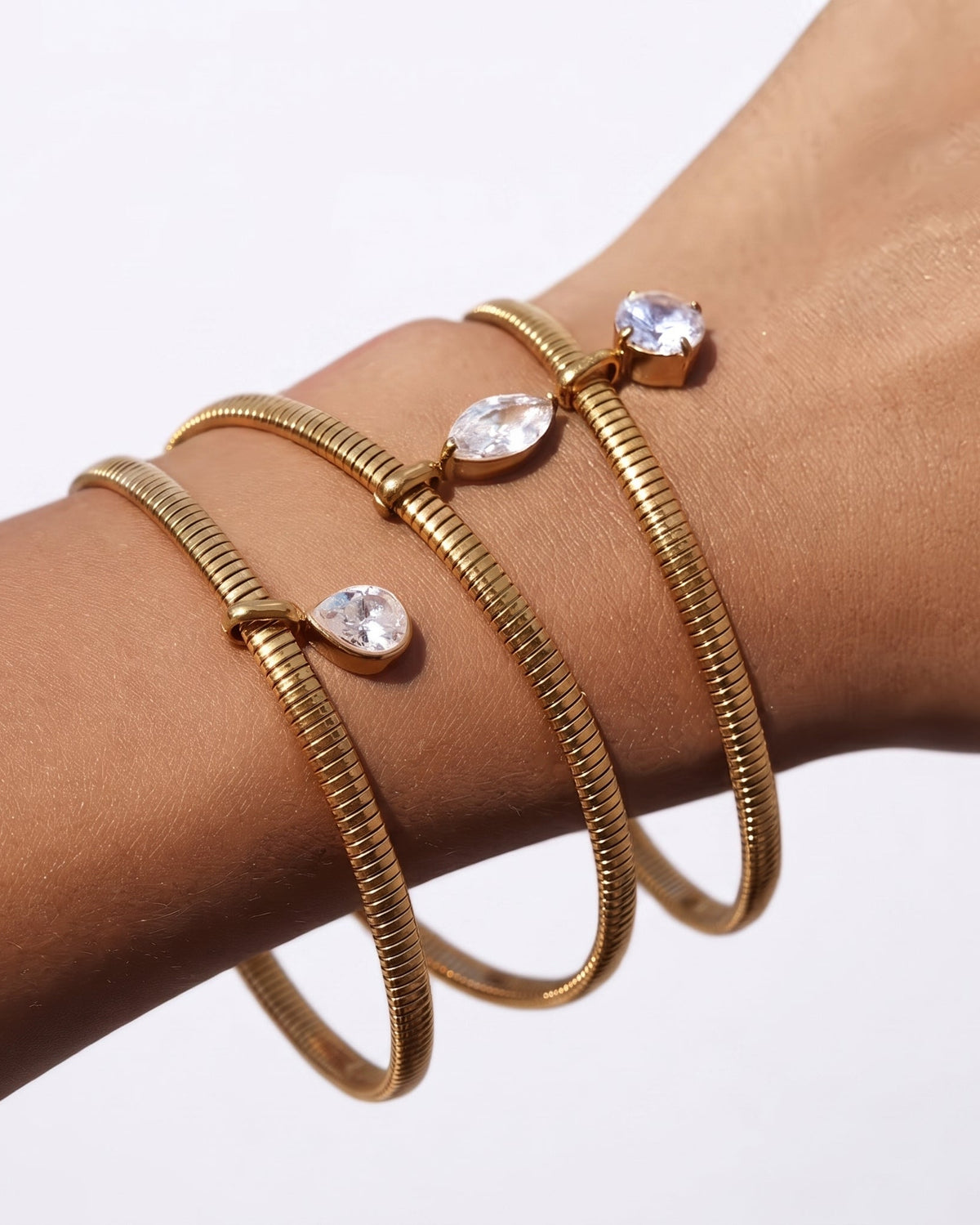 Three gold snake-bangle bracelets with clear teardrop and oval gemstones, shown wrapped on a person's wrist against a plain white background.