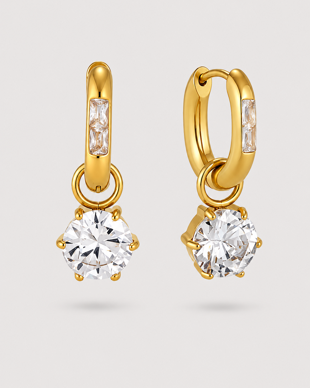 A pair of gold huggie hoop earrings with rectangular baguette-cut clear stones in the upper loop and a large round clear gemstone in a prong setting hanging below, shown on a light gray background.
