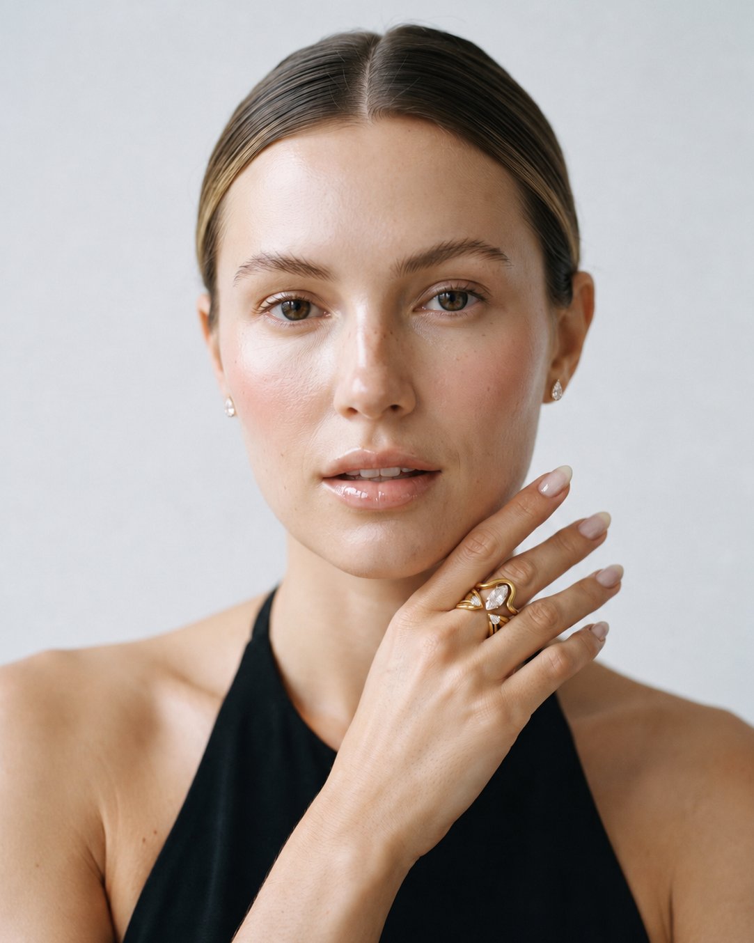 A close-up portrait of a woman wearing a large gold ring with a heart-shaped clear gemstone on her finger, set against a plain light gray background.