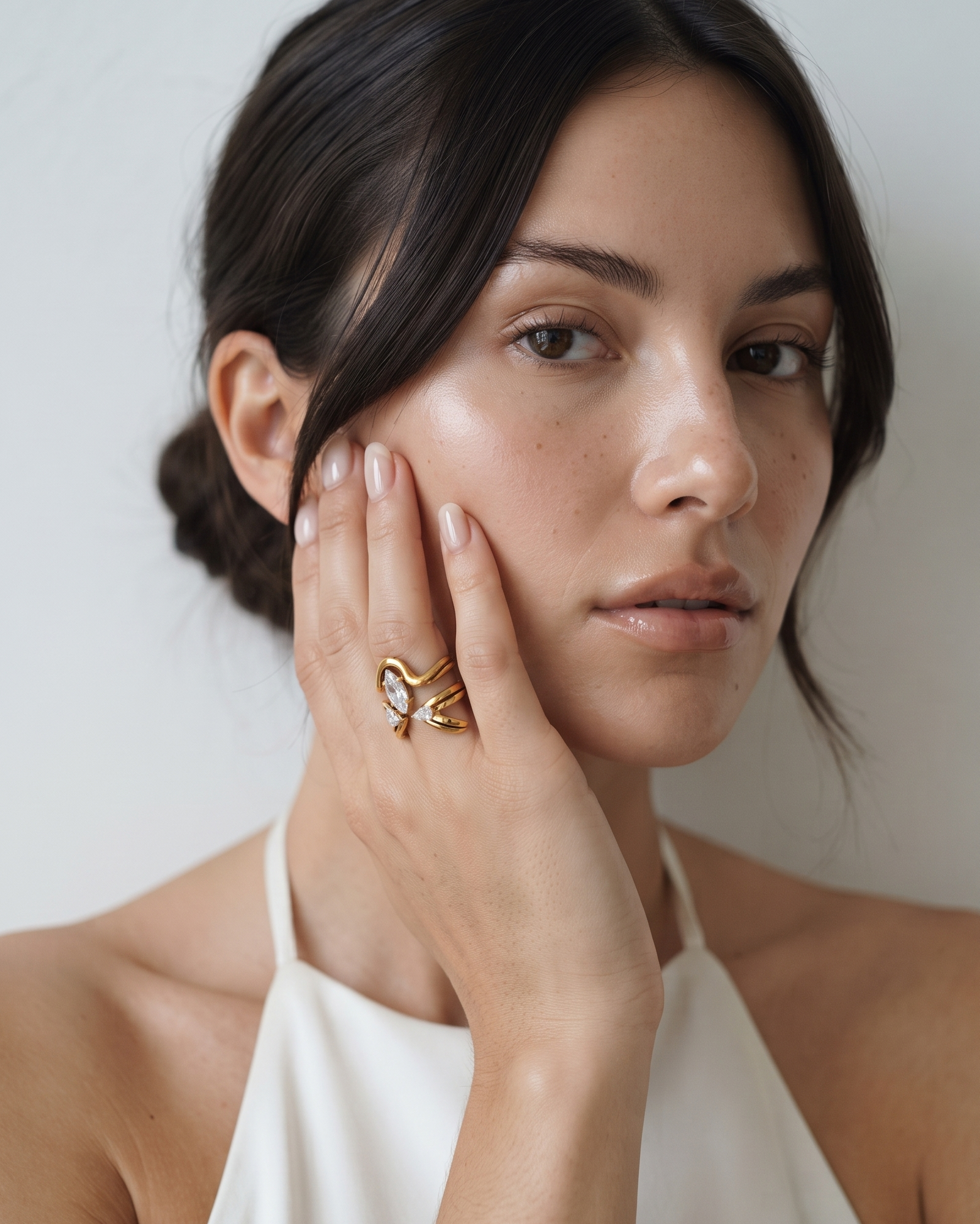 A close-up portrait of a woman with dark hair in a low bun, wearing a white halter top and a gold ring with a white stone on her finger, set against a plain light gray wall.