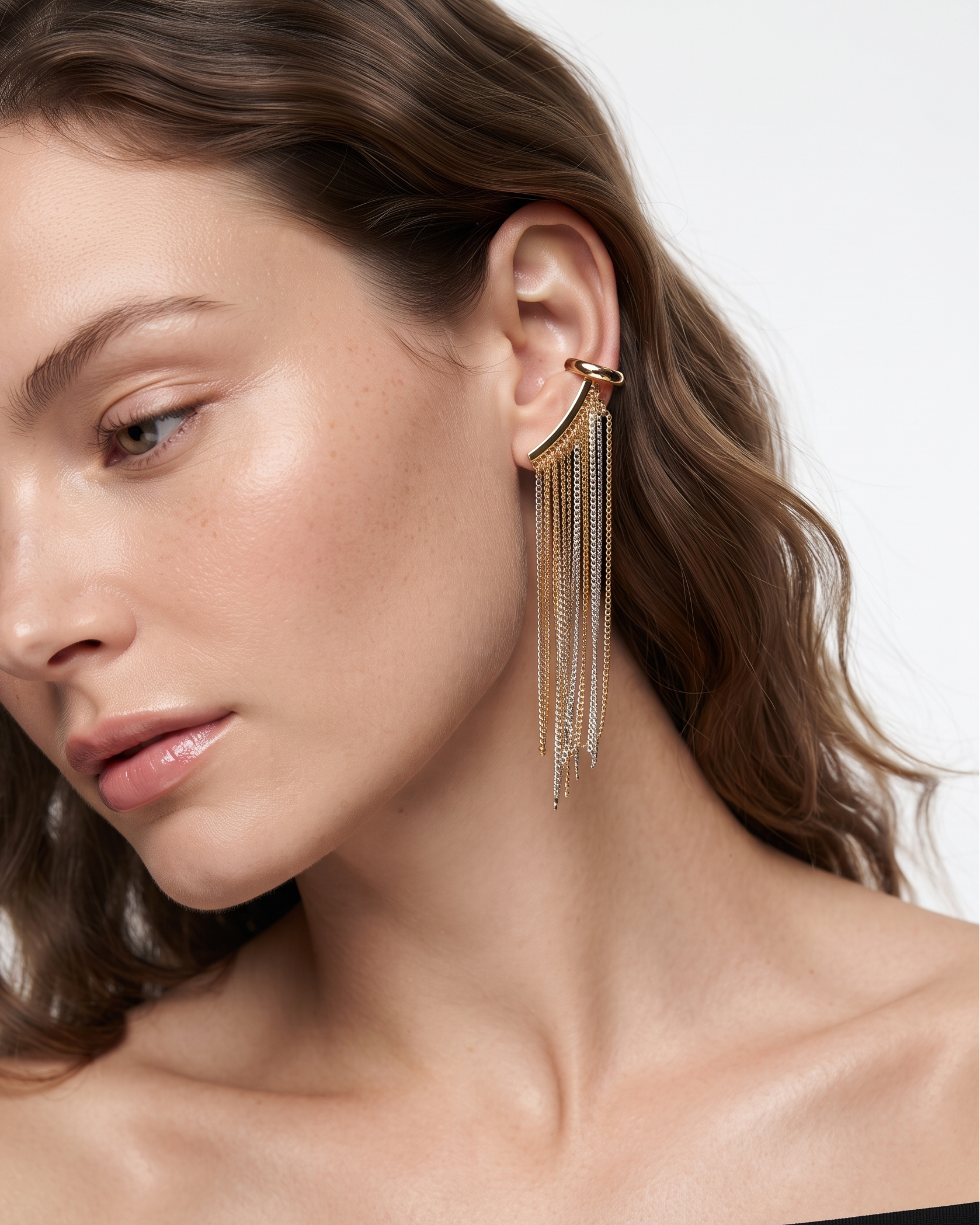 A close-up of a woman wearing a gold-toned curved hoop earring with delicate dangling chain fringe, shown against a clean light background.