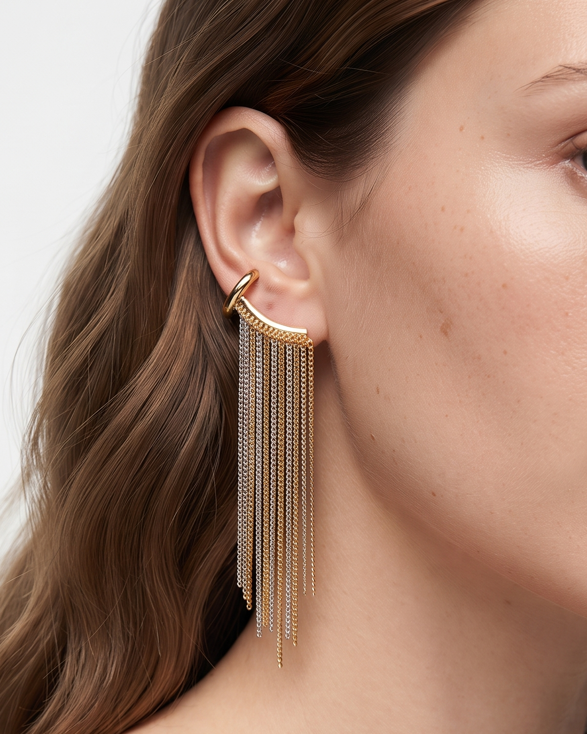 A close-up of a woman wearing a gold crescent-shaped hoop earring with long dangling chain fringe, shown against a clean light background.