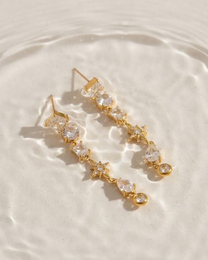 A pair of gold drop earrings with clear faceted gemstones, arranged on a textured white surface with a soft water ripple effect.