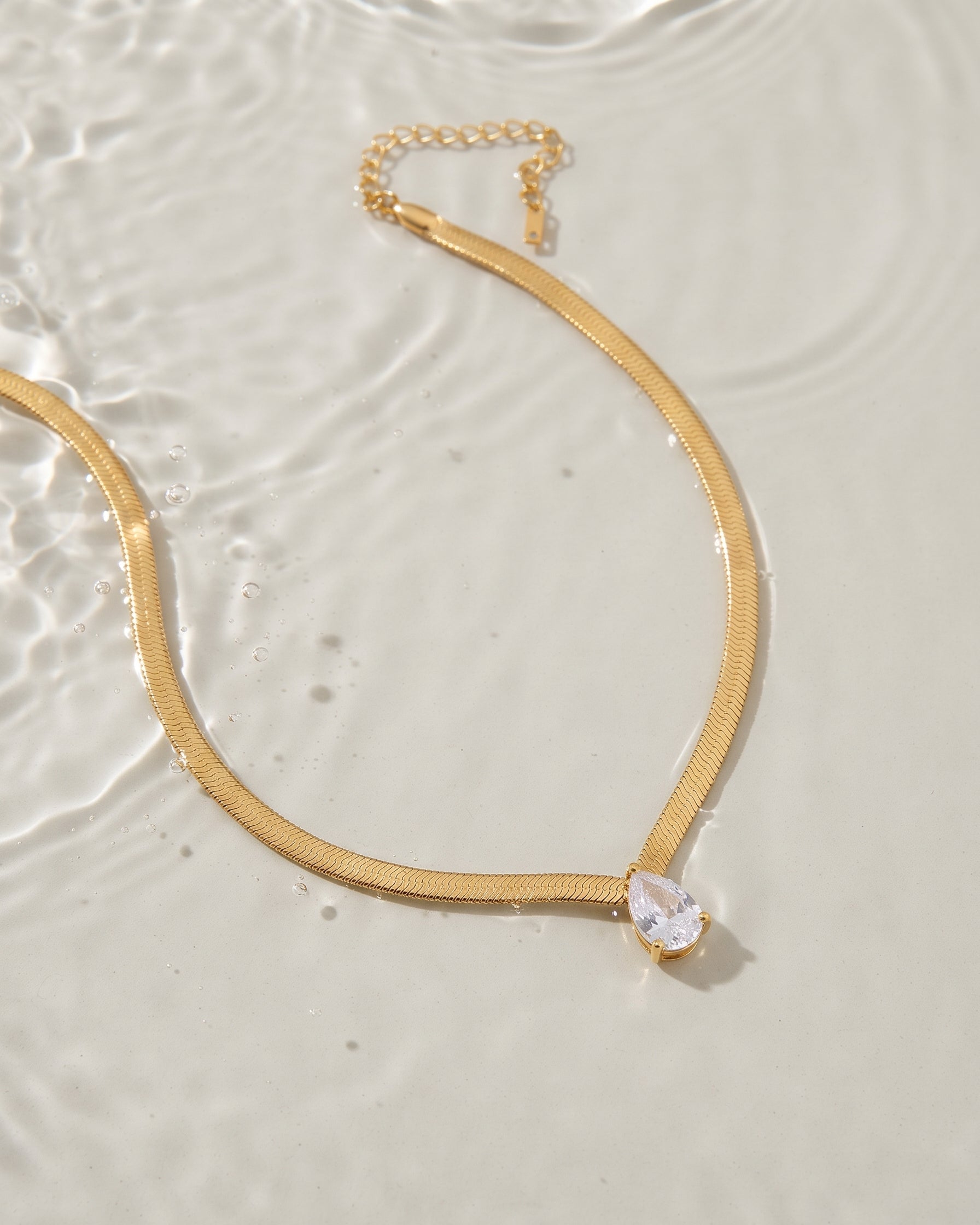 A gold snake chain necklace with a teardrop-shaped clear crystal pendant, laid flat on a light beige surface with water ripples in the background.