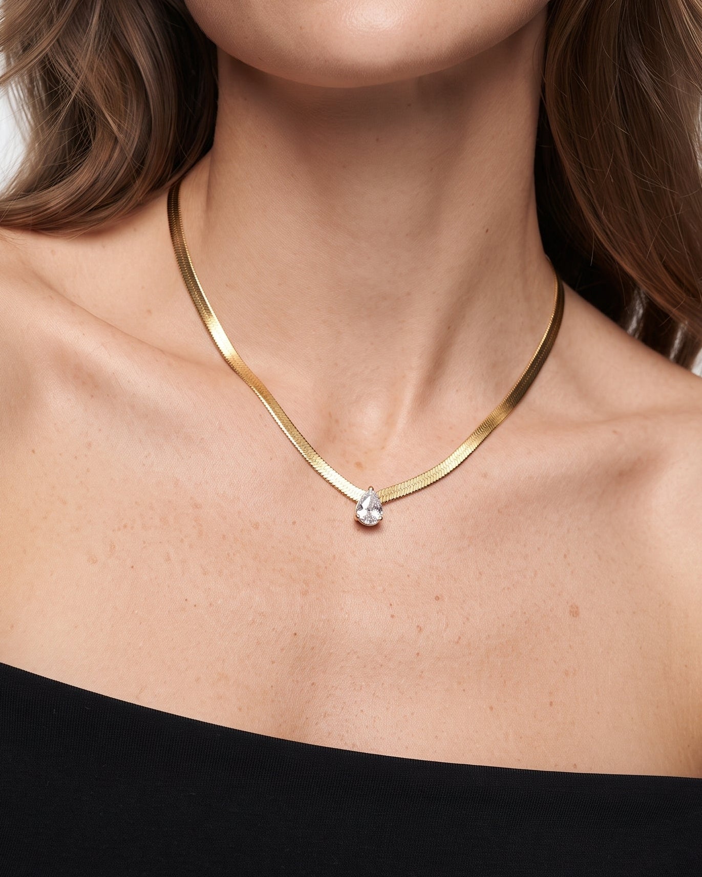 A close-up of a woman wearing a delicate gold necklace with a small clear teardrop gemstone pendant, styled with a black off-the-shoulder top against a light gray background.