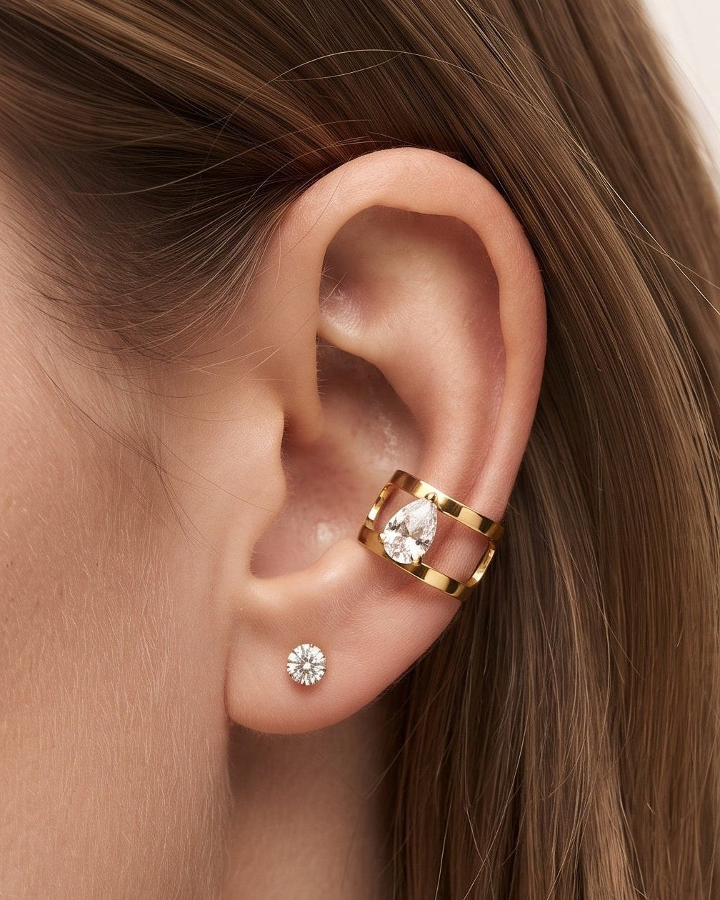 A close-up of a model's ear showcasing a gold ear cuff with a clear crystal center, alongside a small silver stud earring, against a soft beige background.