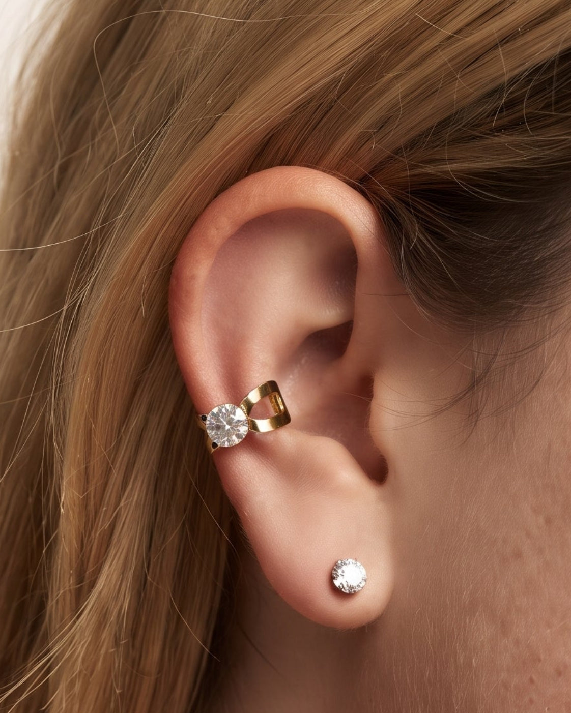 Close-up of a woman's ear wearing a gold butterfly-shaped ear cuff with a clear gemstone and a small silver stud earring, against a plain light beige background.