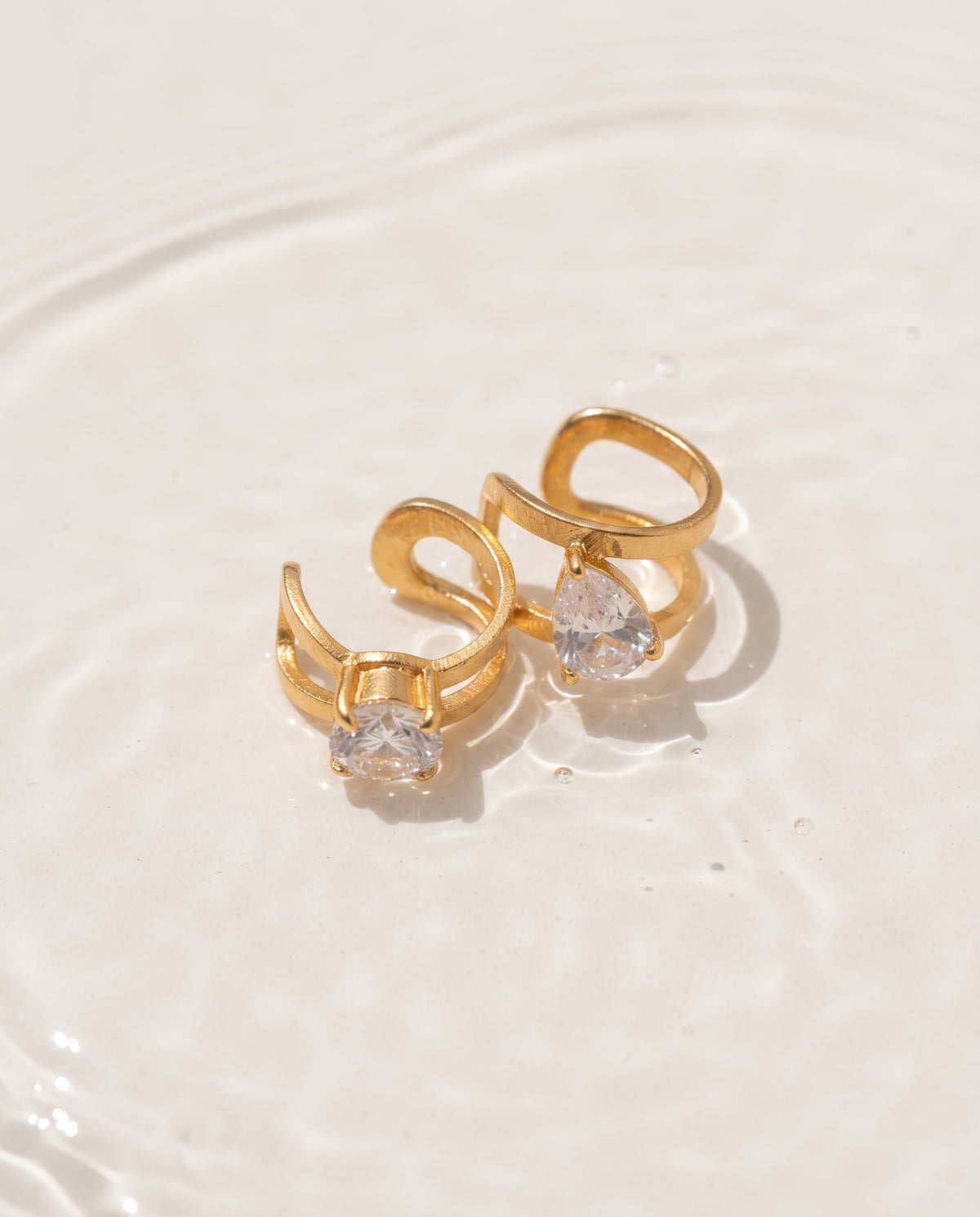A pair of gold-toned ring earrings with clear teardrop-cut gemstones, resting on a white surface with subtle water ripples.