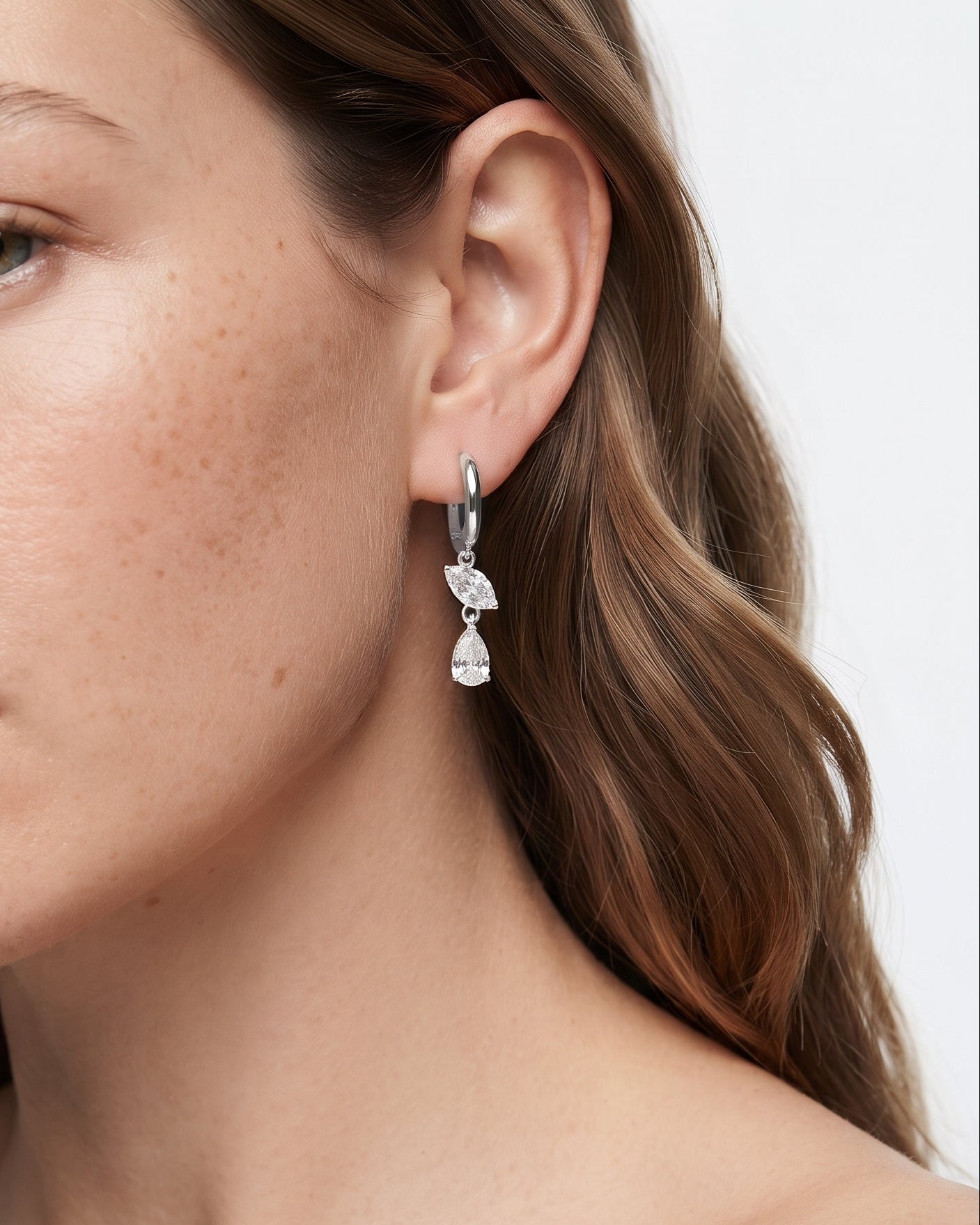 A close-up of a woman wearing a silver huggie hoop earring with three dangling clear crystal pendants, set against a clean white background.