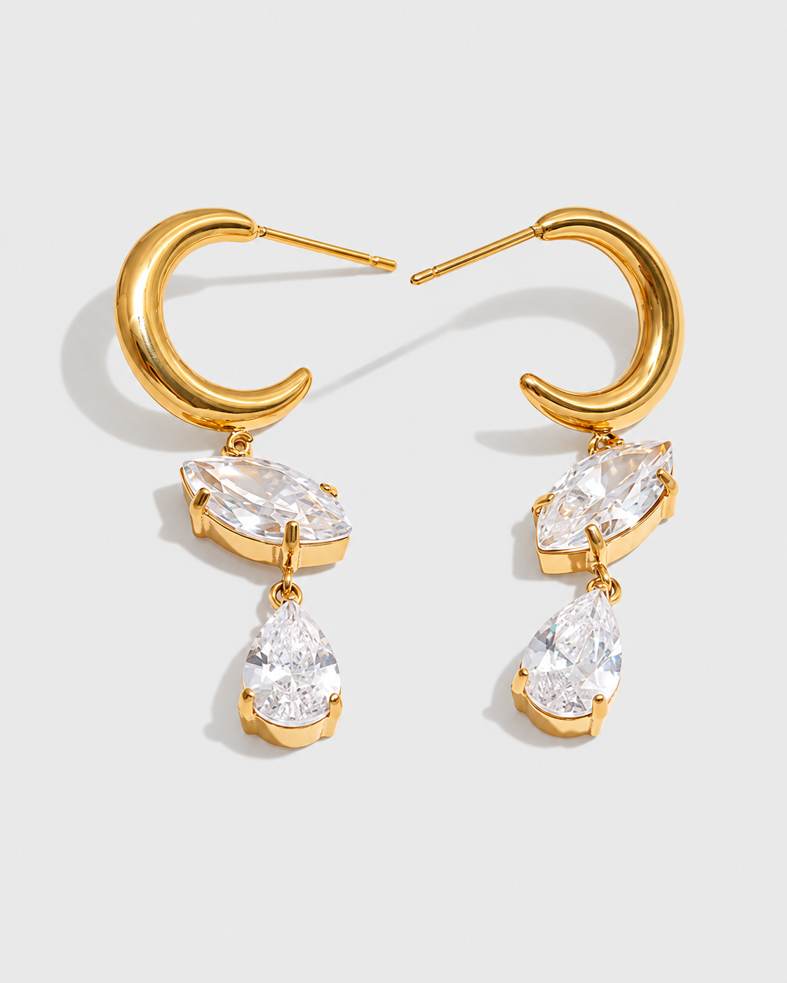 A pair of gold hoop earrings with dangling clear marquise and teardrop gemstones, photographed on a clean light gray background.
