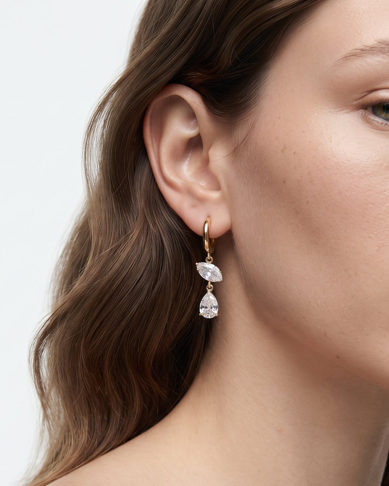 A close-up of a woman wearing a delicate gold huggie hoop earring with two cascading pear-cut clear gemstones, set against a clean white background.