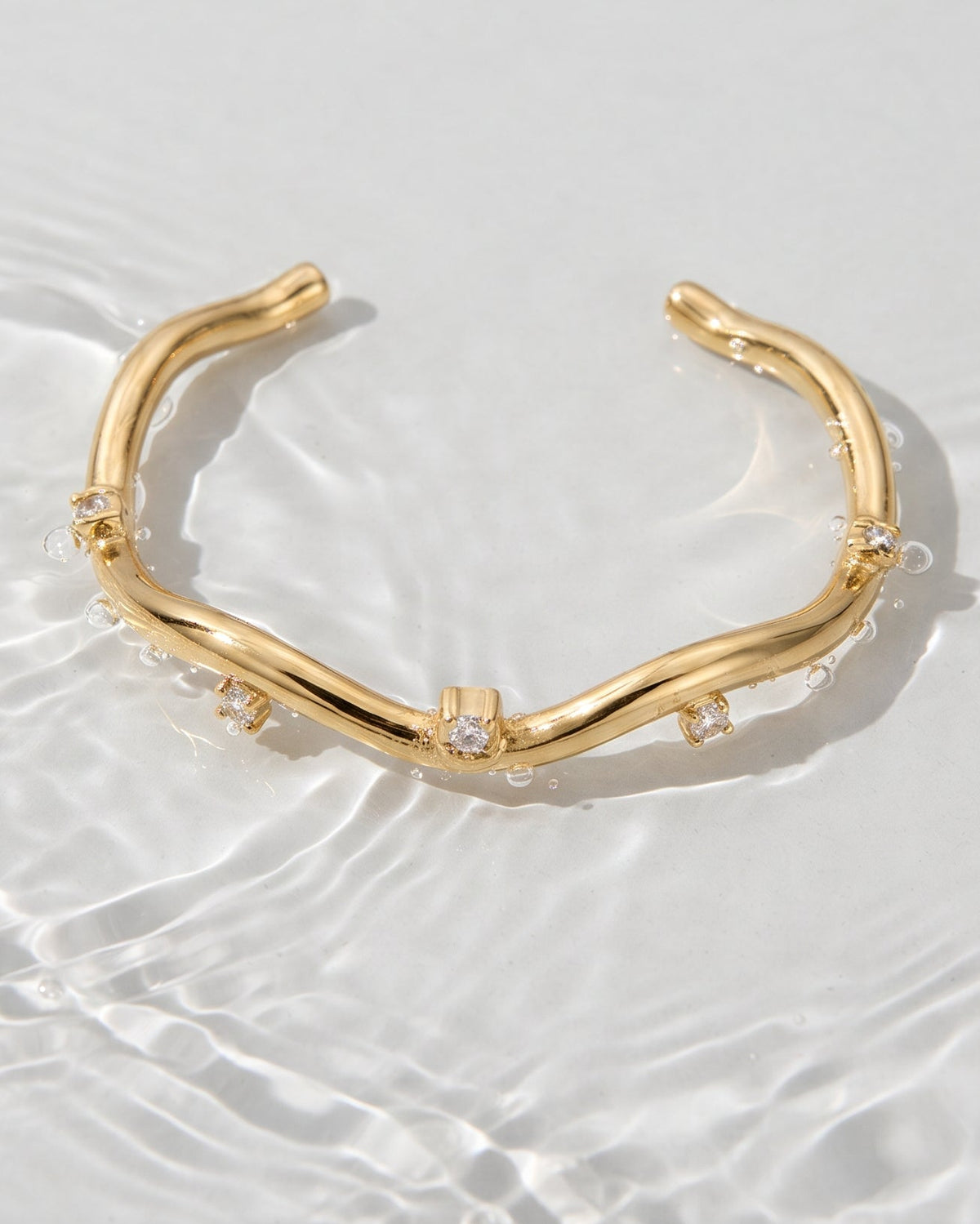 A gold open cuff bracelet with organic, wavy branches and small clear crystal accents, displayed on a white surface with water ripples.