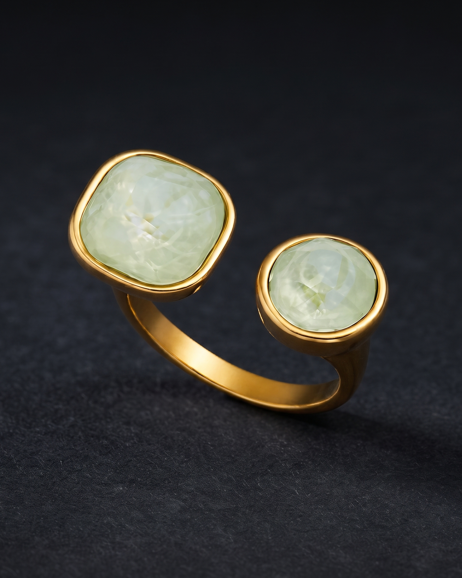 A gold-toned ring with two round bezel-set pale green stones, photographed on a dark textured background.