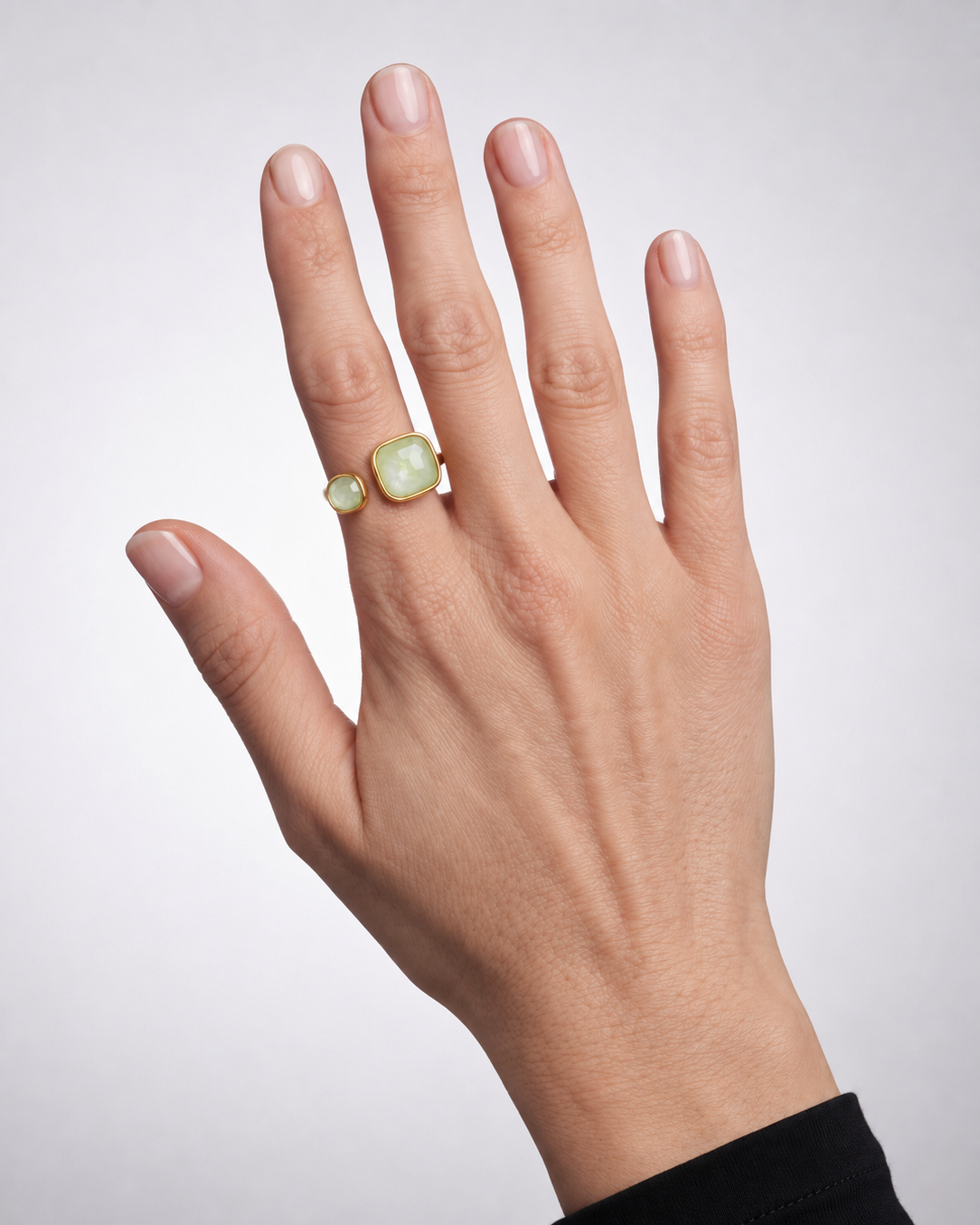 A close-up of a hand wearing a gold ring with a large, rounded, pale green cabochon gemstone and a smaller matching green gemstone on the ring finger, set against a light gray background.