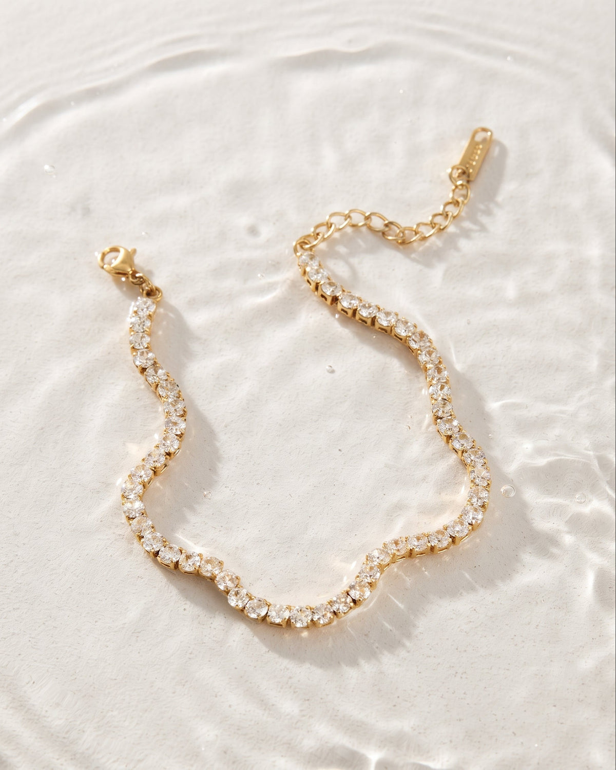 A delicate gold chain anklet embellished with clear rhinestones, arranged in a wavy line on a textured white surface with soft water ripples.