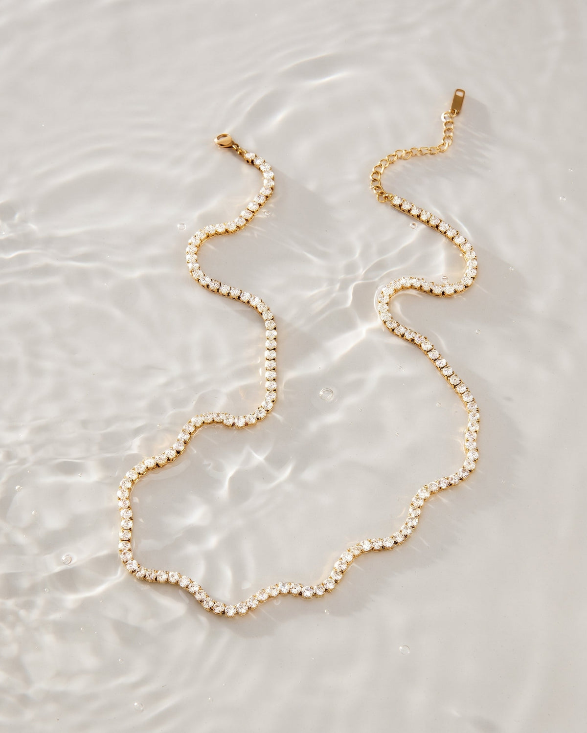 A delicate gold chain necklace adorned with small, clear faceted beads, arranged in a wavy line on a white, water-textured surface.