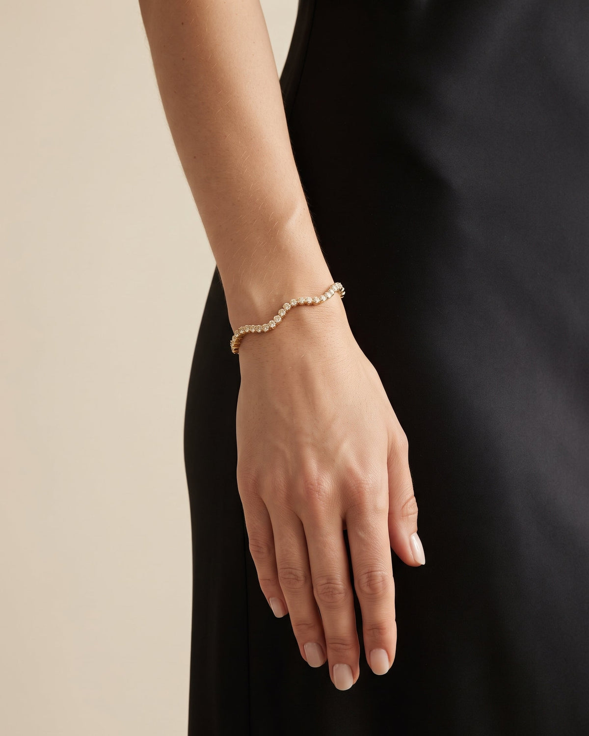 A delicate gold chain bracelet with small clear stones worn on a woman's wrist, shown against a black dress and a warm beige background.