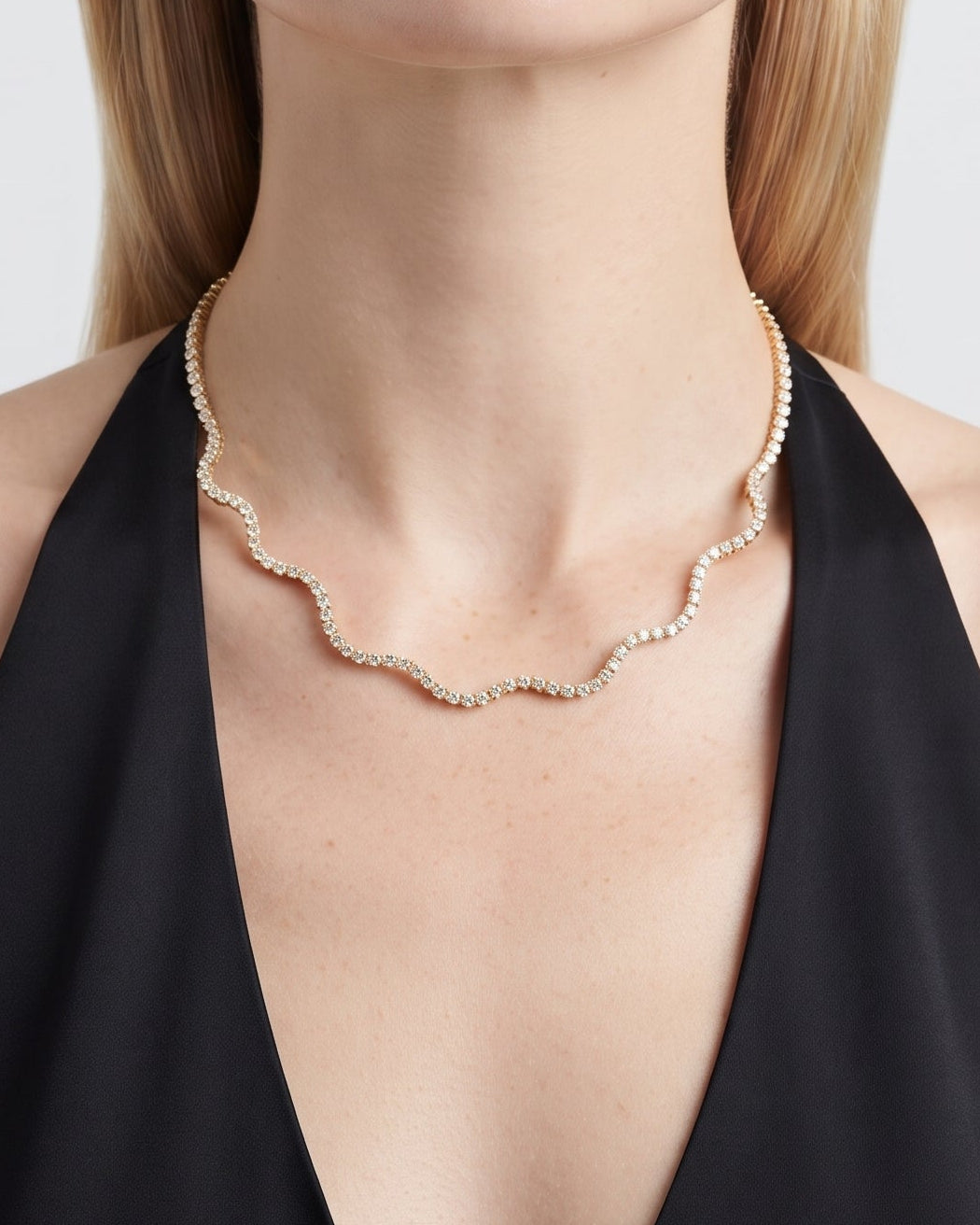 A close-up of a blonde model wearing a delicate gold scalloped chain necklace with a scalloped pearl collar, paired with a black halter top against a light gray background.