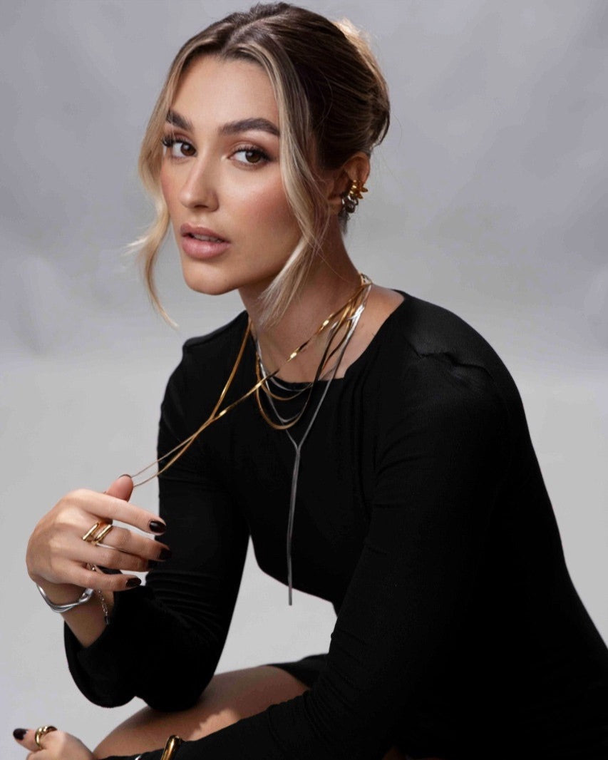 Woman wearing multiple necklaces and holding a pair of sunglasses against a gray background