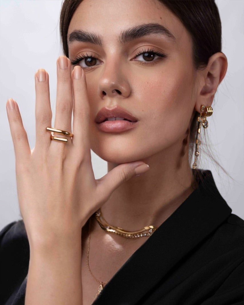 Woman wearing gold jewelry including a ring, necklace, and earrings against a neutral background
