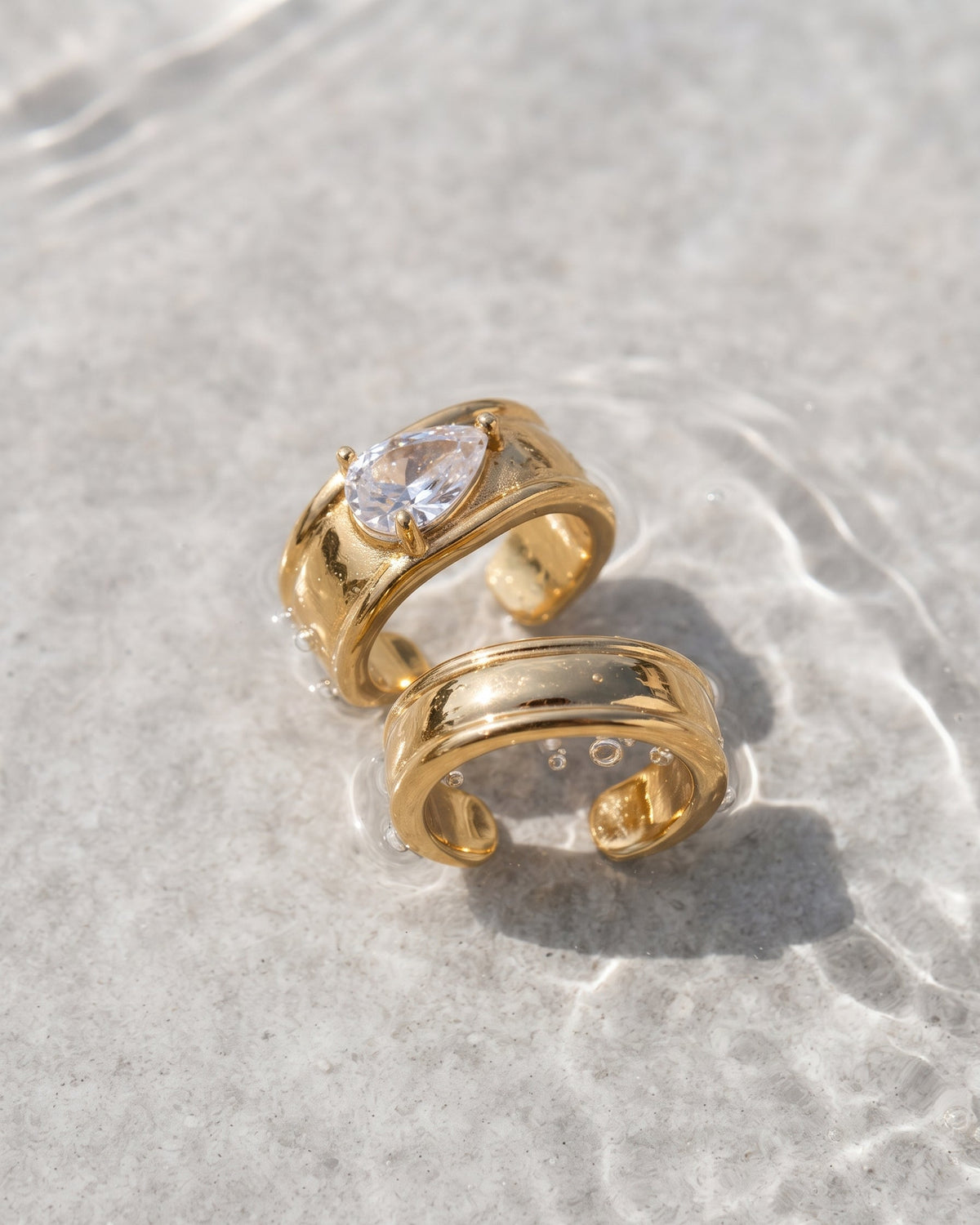 Two gold-toned open bangle rings resting on shallow water over a light gray stone surface. One ring features a teardrop-shaped clear gemstone, and the other is plain with a polished finish.