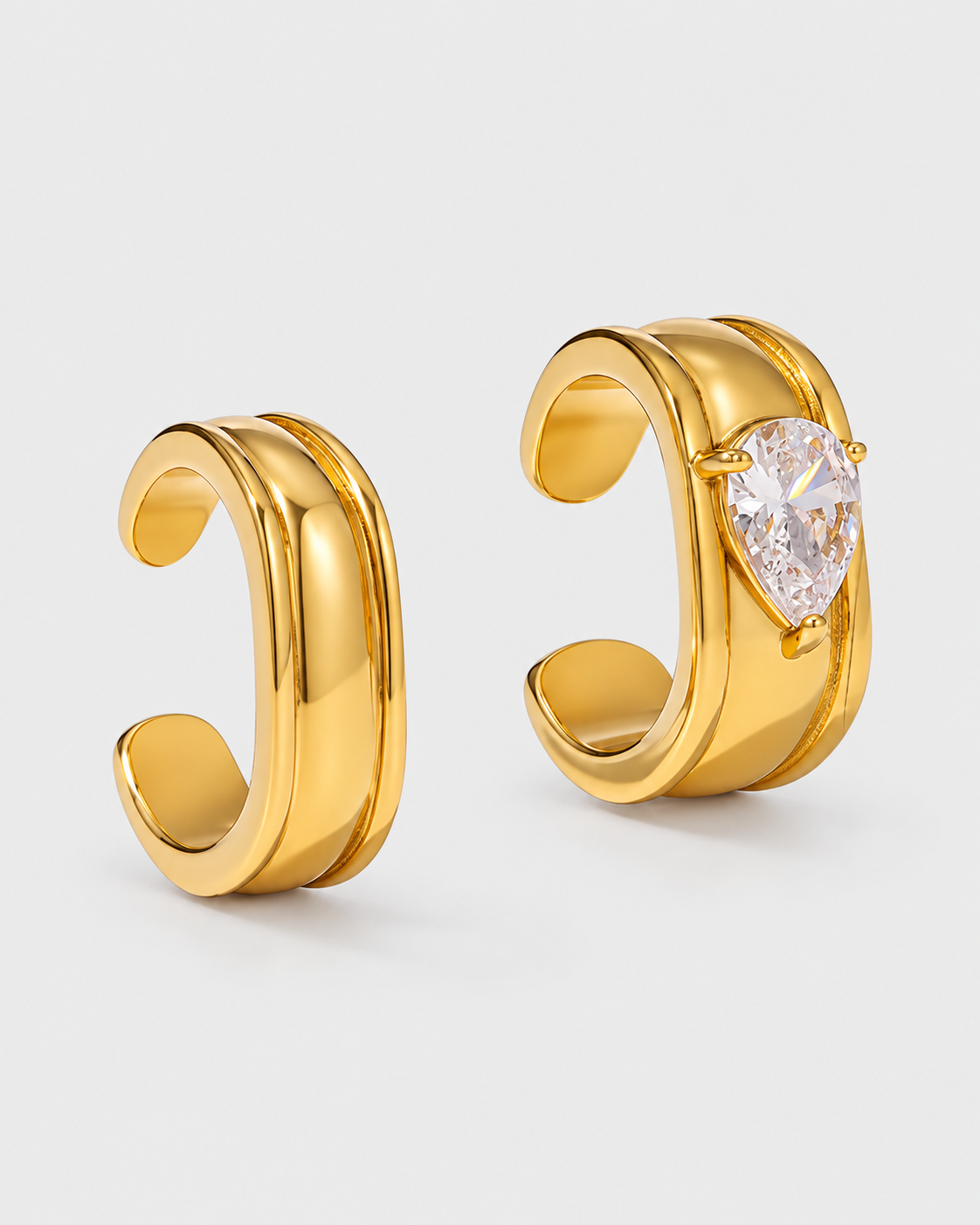 A pair of gold huggie hoop earrings, one plain and one featuring a large pear-cut clear gemstone, set against a light gray background.