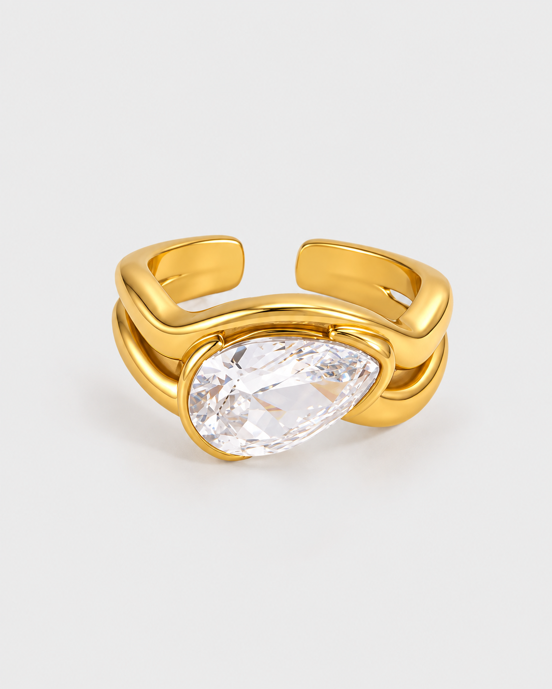 A gold-plated open ring with a large, clear teardrop-shaped gemstone in the center, set against a light gray background.
