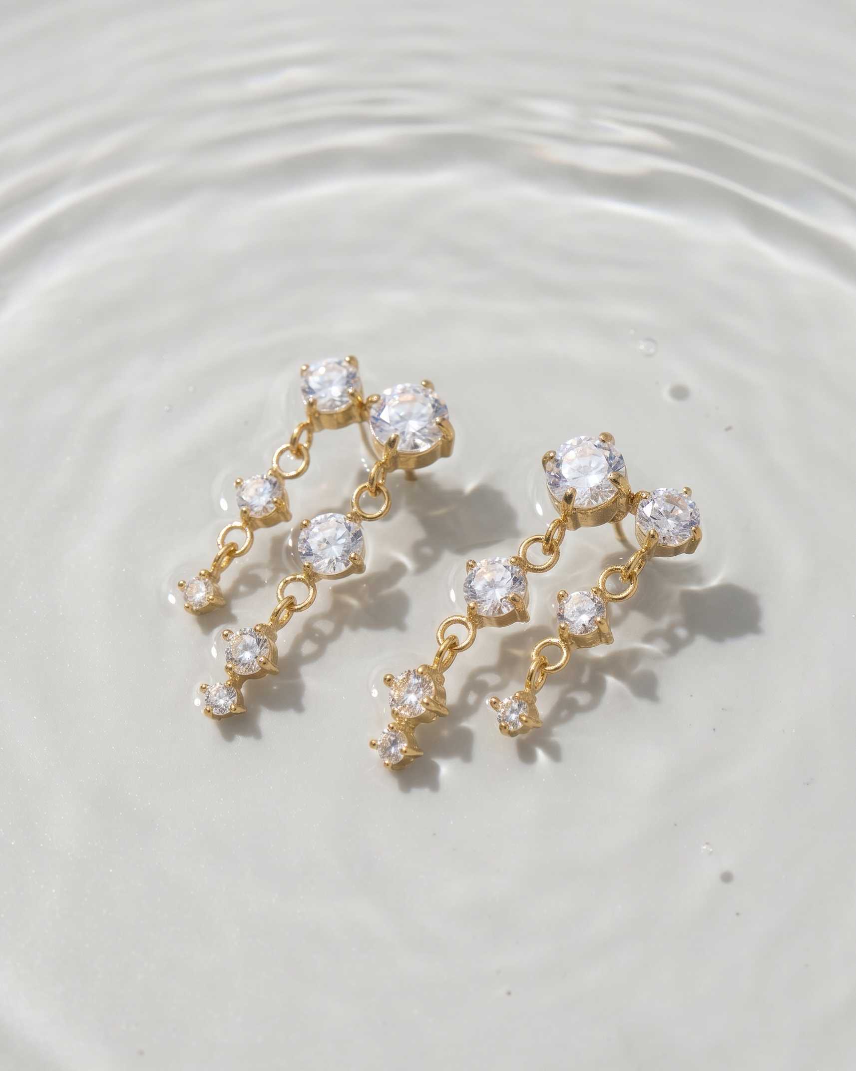 Gold earrings with clear stones on a white surface with water ripples
