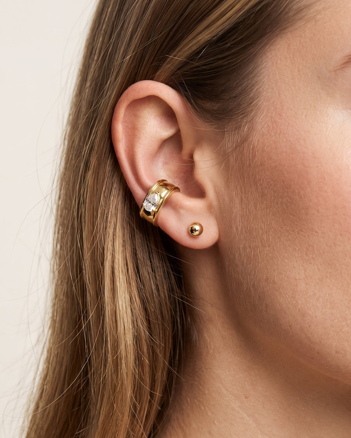 A close-up of a model's ear showcasing a gold huggie hoop earring adorned with small clear crystals, alongside a small gold stud earring, against a soft beige background.