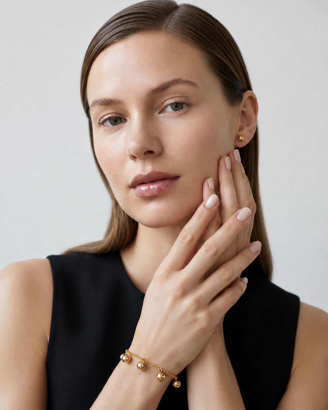A close-up of a woman with brown hair wearing a delicate gold chain bracelet with small round bead charms, posing with her hand near her face against a light gray background.