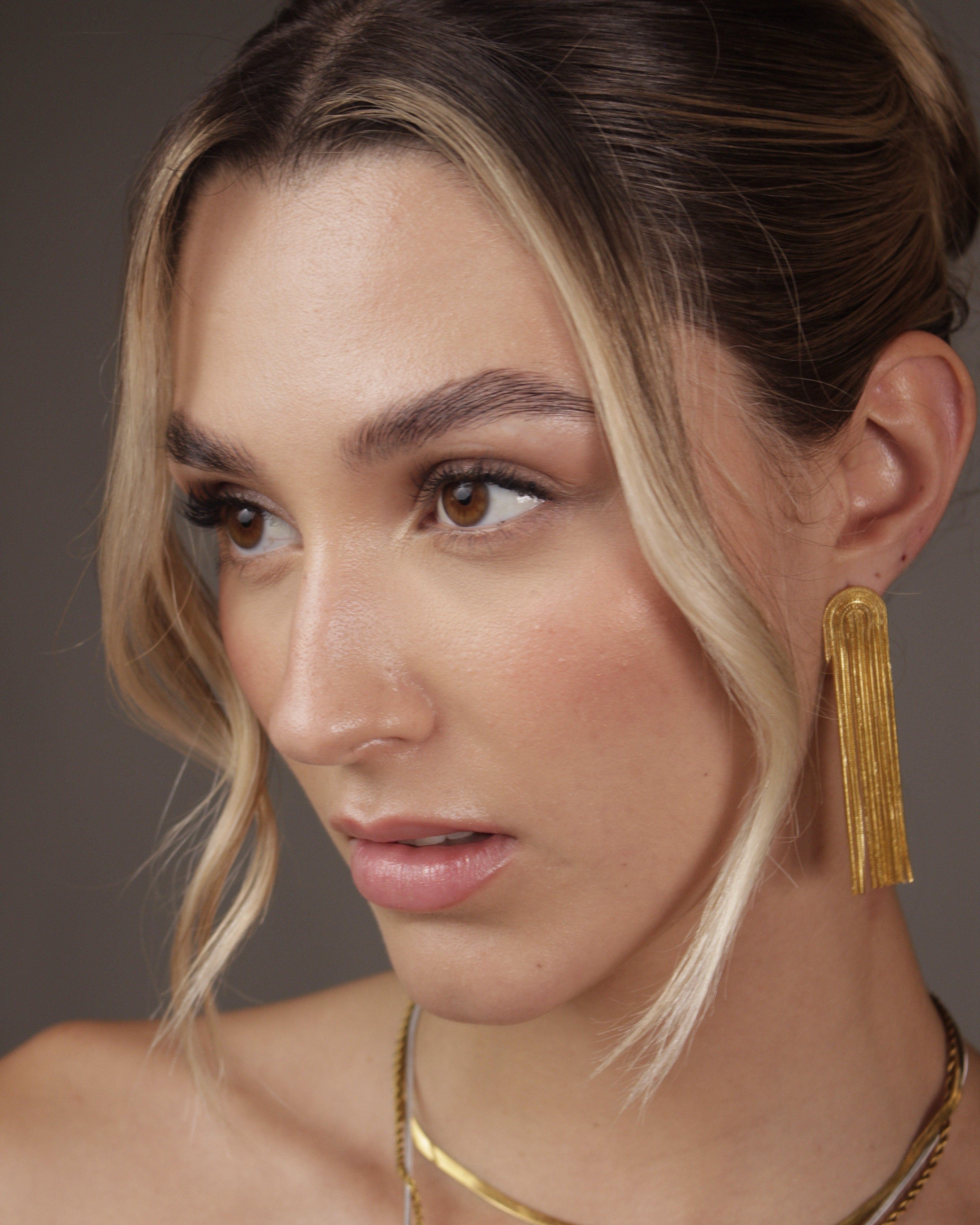 Close-up of a person wearing gold earrings and necklaces against a neutral background