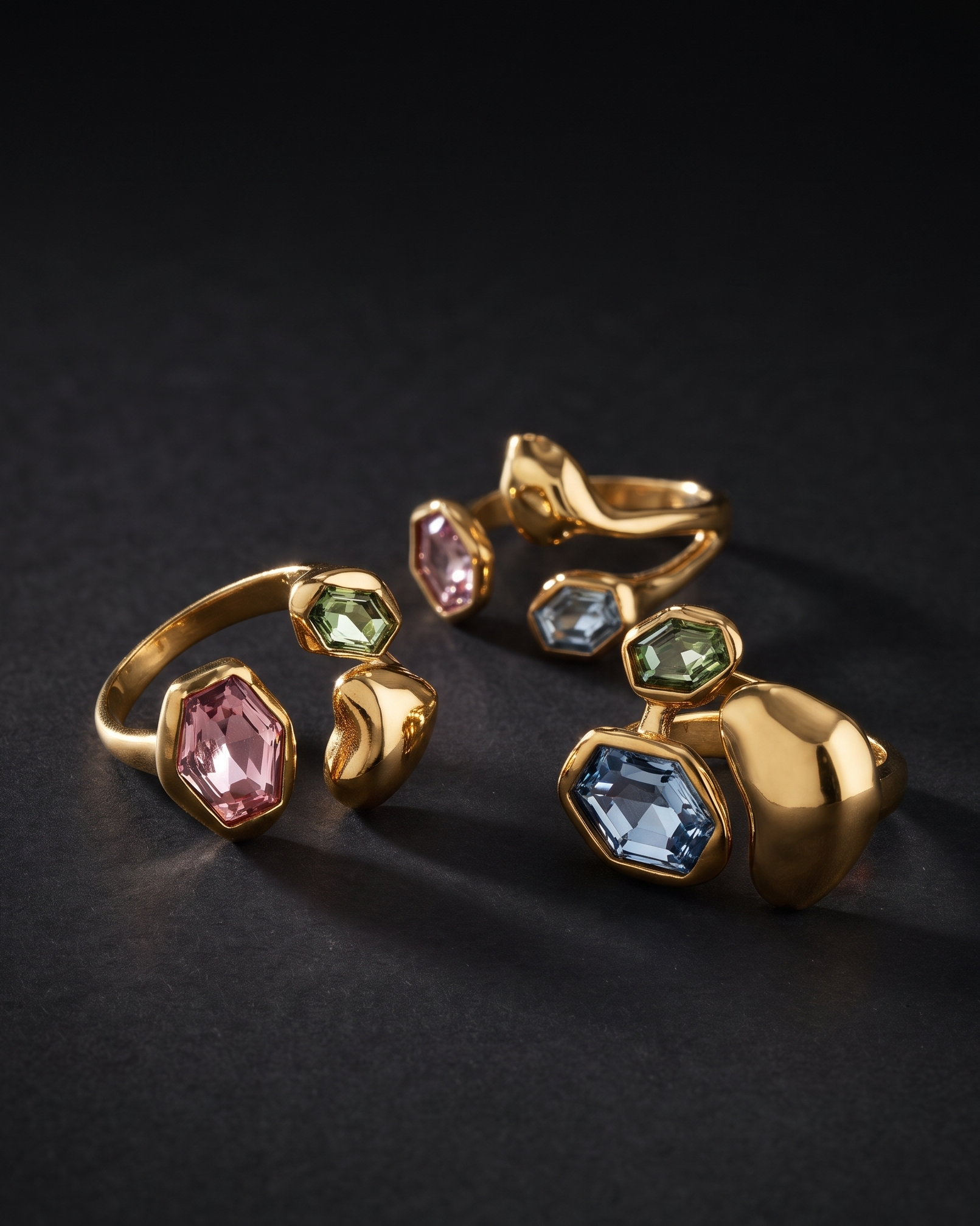 Three gold rings with asymmetrical multicolored gemstone clusters, including pink, green, and blue stones, arranged on a dark textured background.