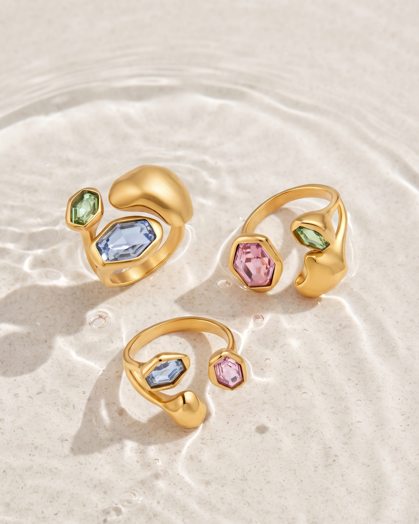 Three gold open ring earrings with abstract organic shapes and colorful gemstones (light blue, pink, and green) arranged on a textured beige surface with water ripples.