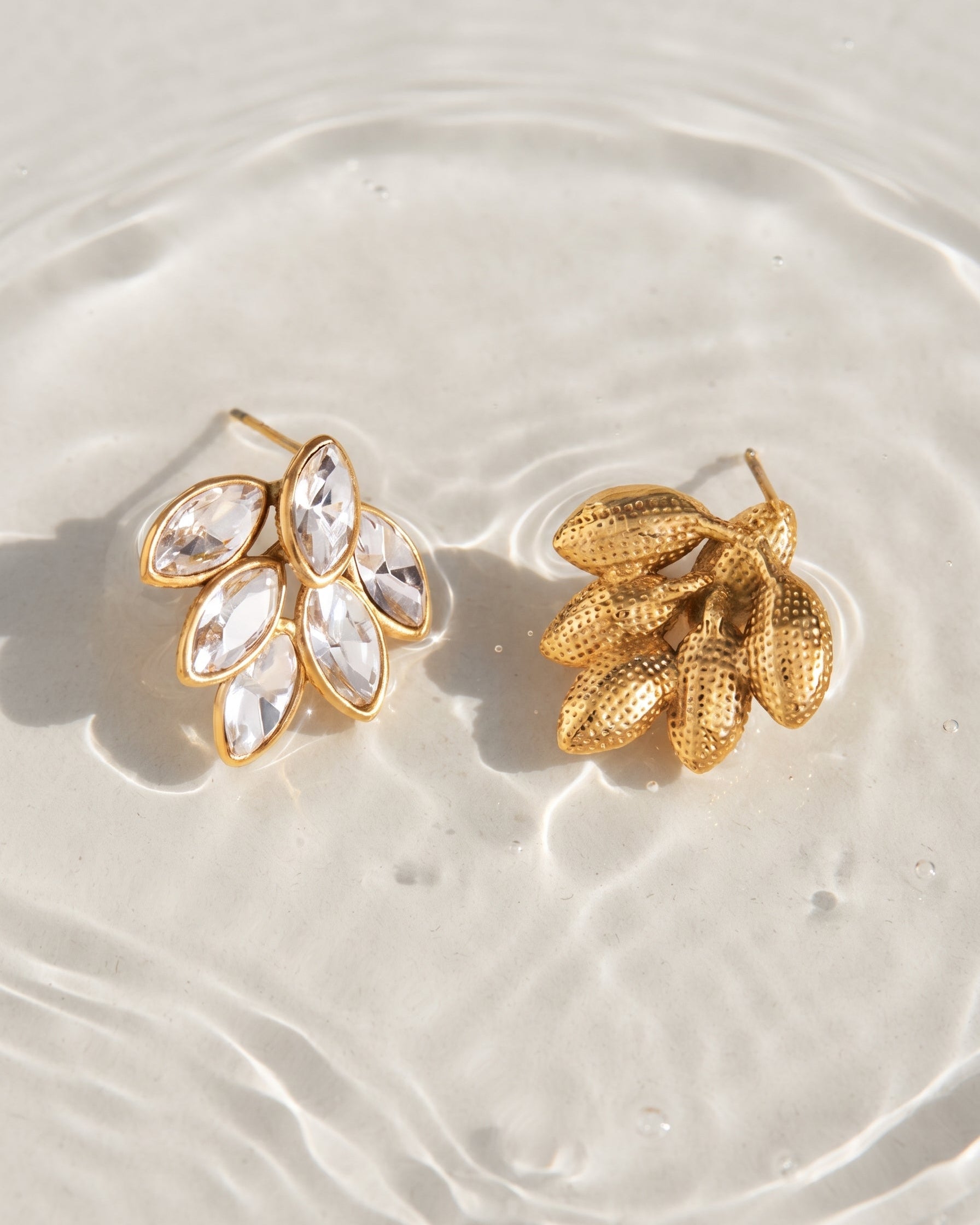 A pair of gold-toned leaf-shaped stud earrings, one with marquise-cut clear crystals and the other with textured gold leaf-like petals, displayed on a light beige surface with water ripples.