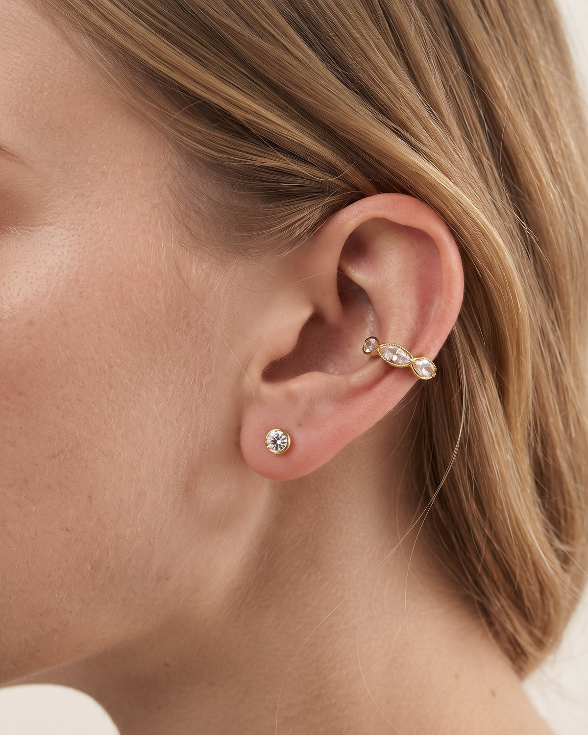 A close-up of a model's ear showcasing a gold ear cuff with a twisted ribbon design and clear crystals, along with a small matching crystal stud earring.