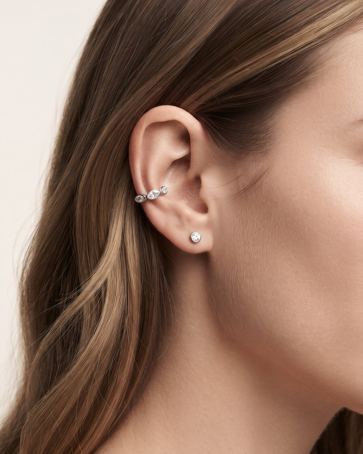A close-up side profile of a woman's ear wearing a silver-toned curved ear cuff with small clear stones and a matching single-stone stud earring, against a soft light beige background.