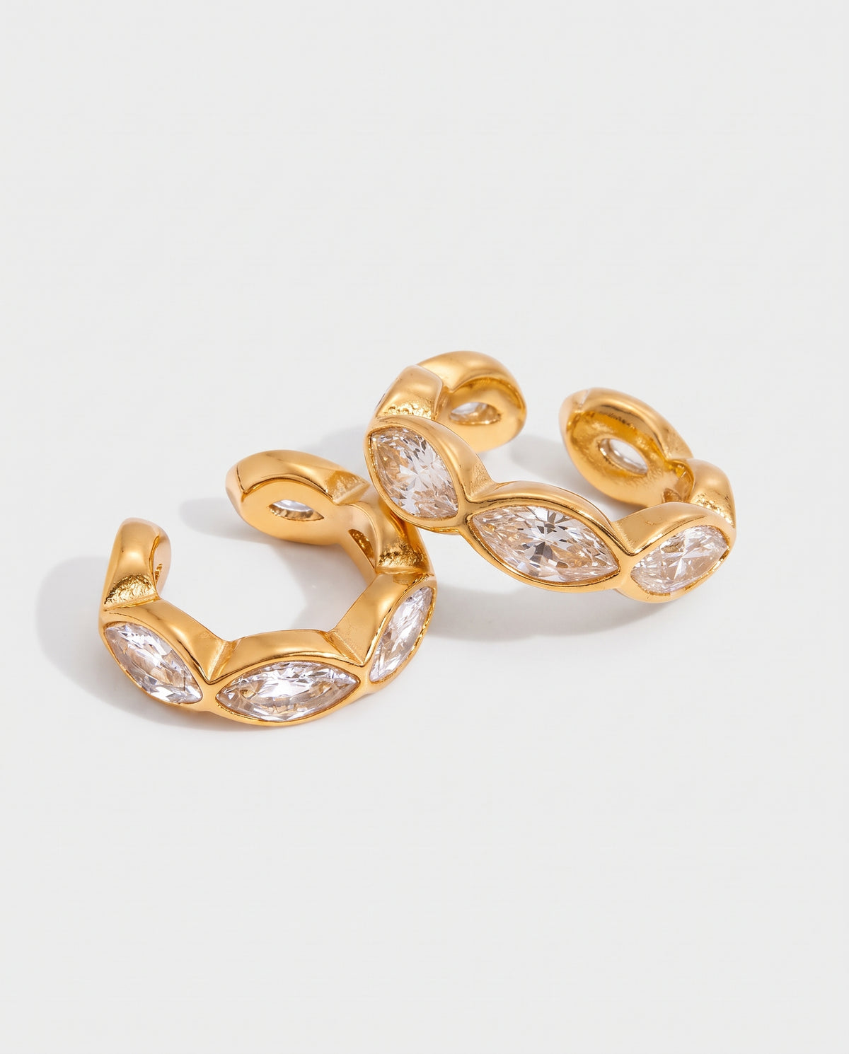 A pair of gold huggie hoop earrings embellished with marquise-cut clear crystals, displayed on a clean light gray background.