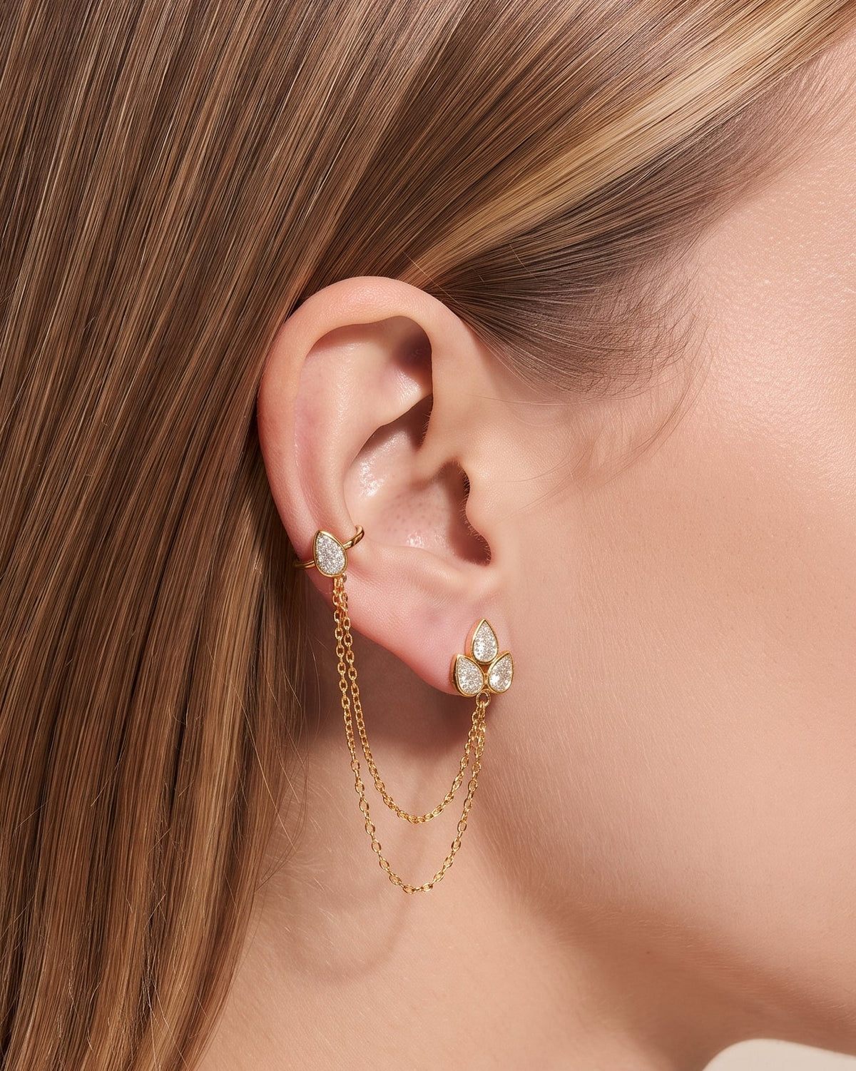 A close-up side profile of a woman wearing a delicate gold ear cuff with small clear stones and two hanging chain drops, against a soft beige background.
