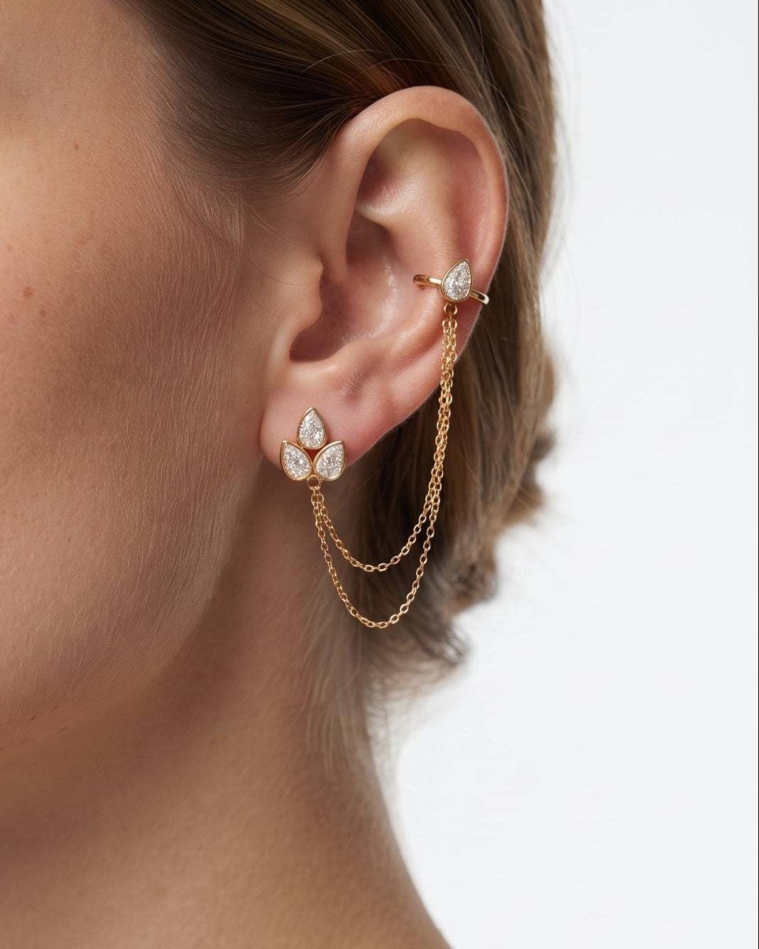 A close-up of a model's ear wearing a gold ear cuff with a teardrop-shaped crystal cluster and two draped chain loops, paired with a small gold stud earring.