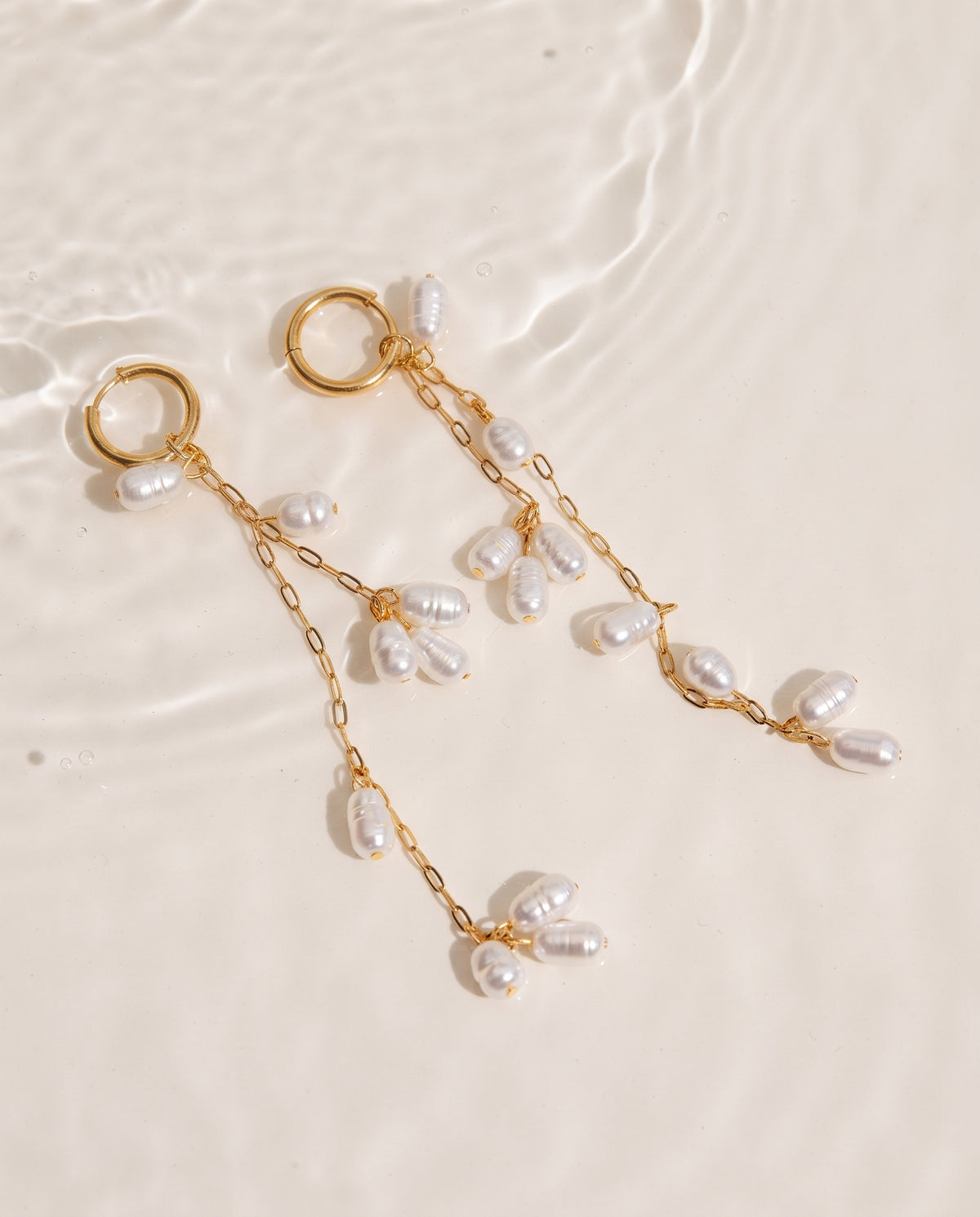 A pair of gold dangle earrings with baroque pearls, displayed on a light beige textured surface with water ripples in the background.