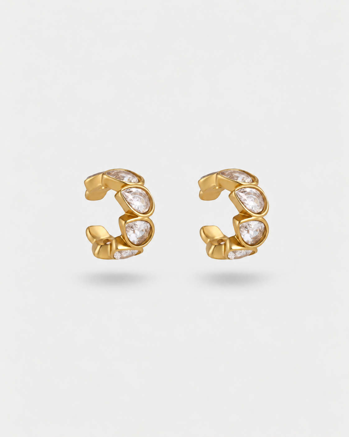 A pair of gold huggie hoop earrings with curved rows of clear marquise-cut gemstones, shown on a light gray background.