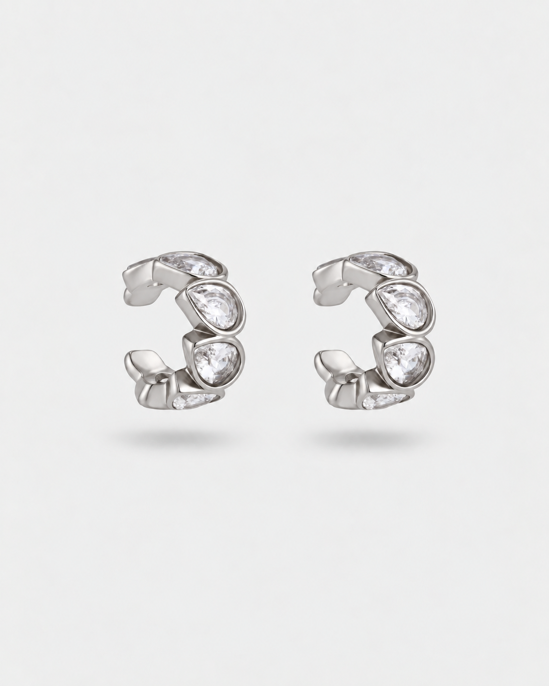 A pair of silver huggie hoop earrings with clear teardrop-shaped crystals, displayed on a light gray background.
