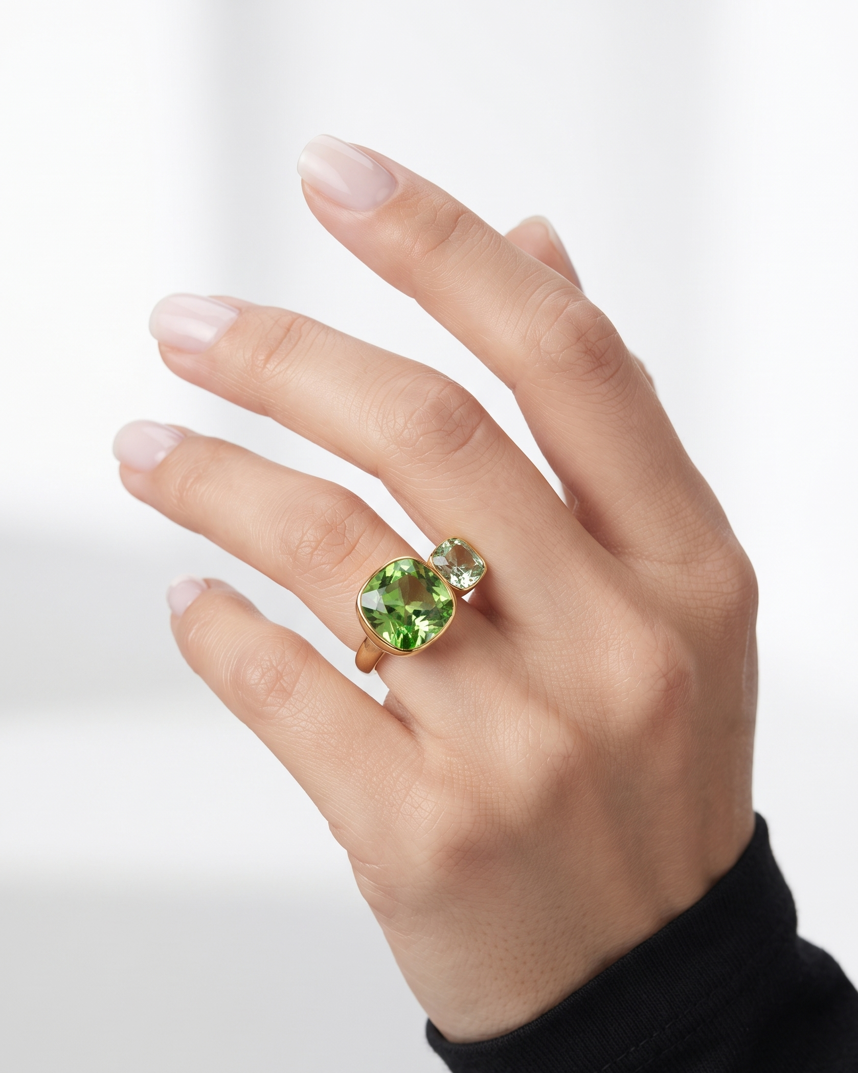 A close-up of a hand wearing a gold ring with a large green heart-shaped gemstone and a smaller clear gemstone beside it, set against a soft white background.